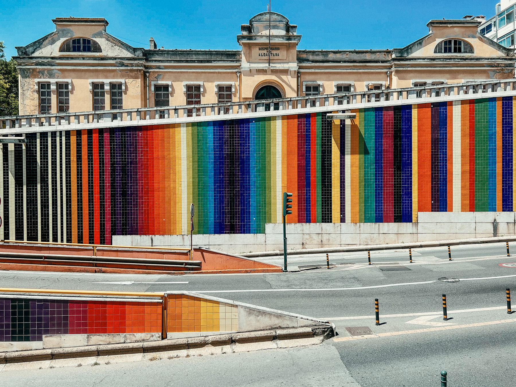 A grandiose building stands behind a very colorful tile wall, overlooking a road, with no cars going by.