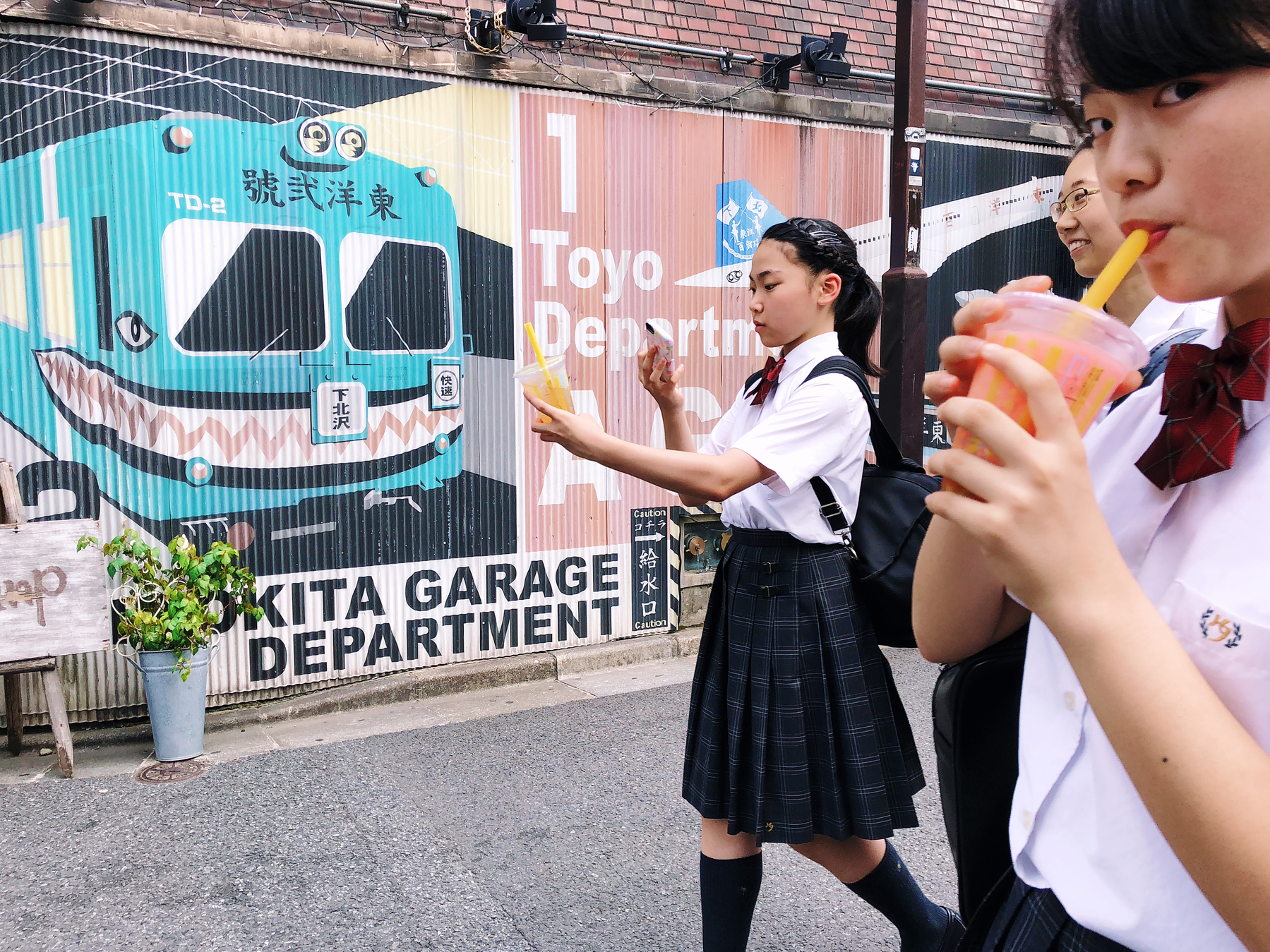 a group of girls walks into the frame from the right side. One is shooting a cup of juice she’s carrying with her cellphone. In the back a train is painted on a wall, Okita Garage Department is written below that