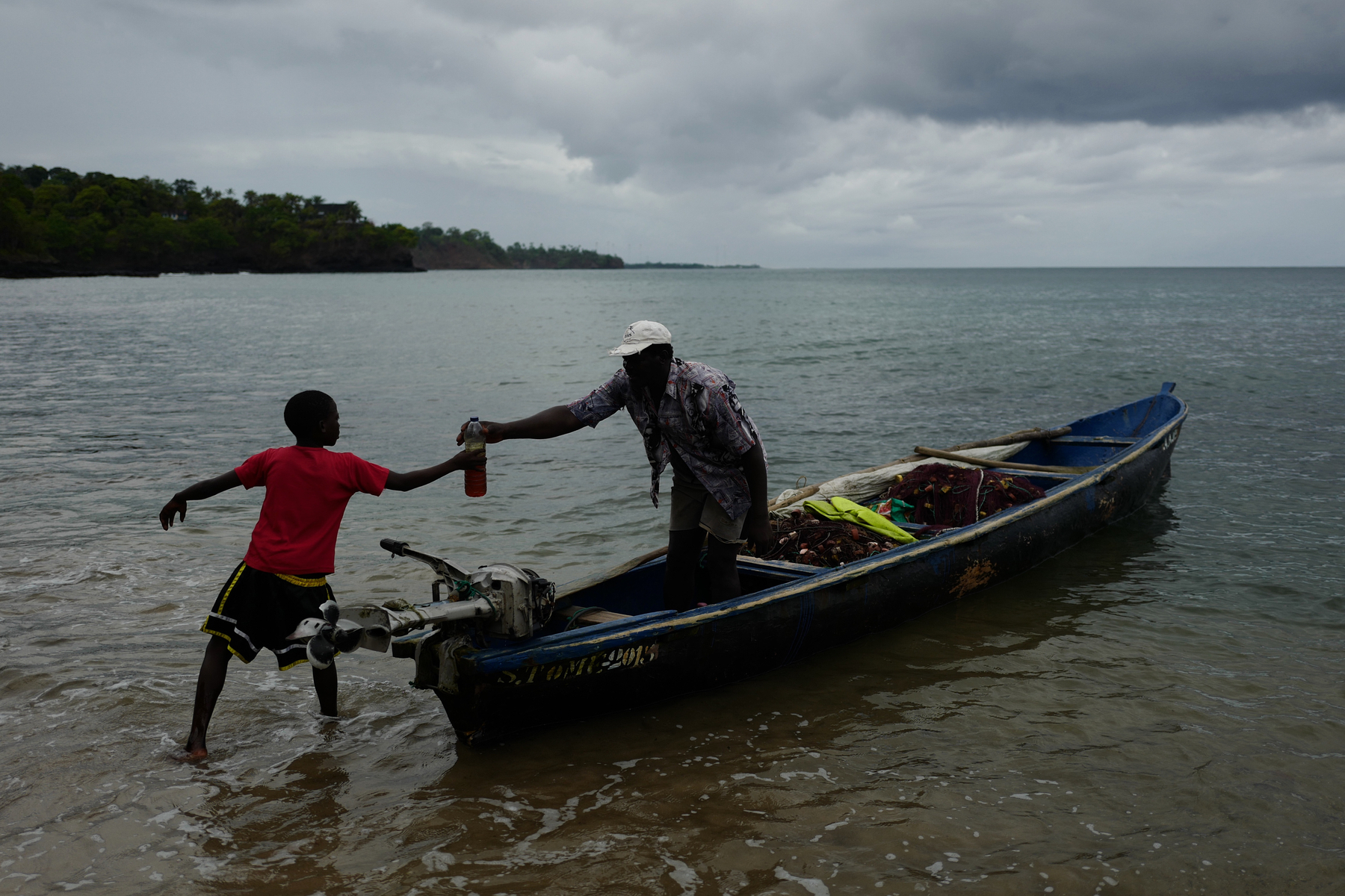A man hands his son a bottle of petrol, while standing on a canoe, in the water. Late afternoon, cloudy skies. 