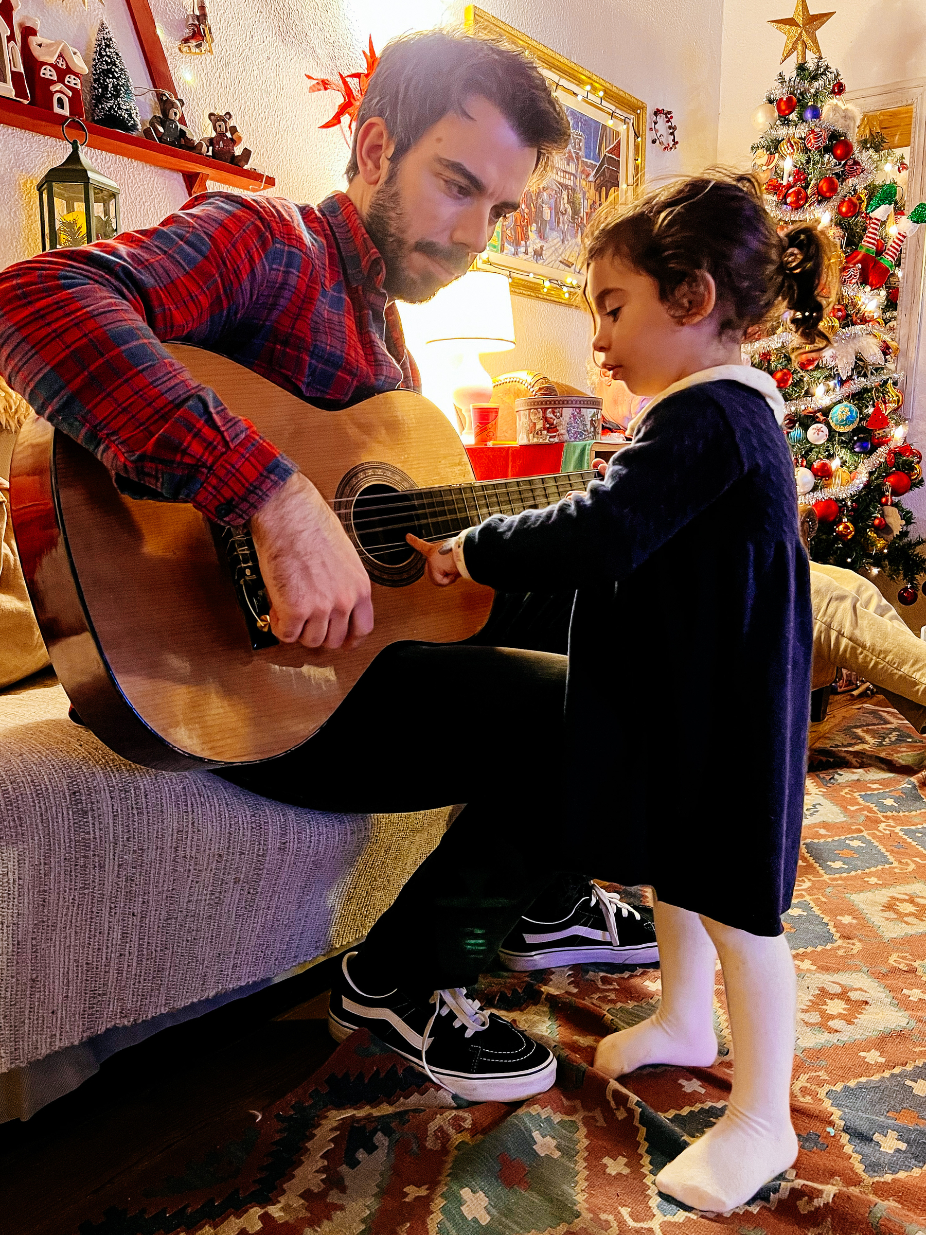 A man holds a guitar while a toddler girls uses her finger to play. The room is fully decked out in Christmas decorations.