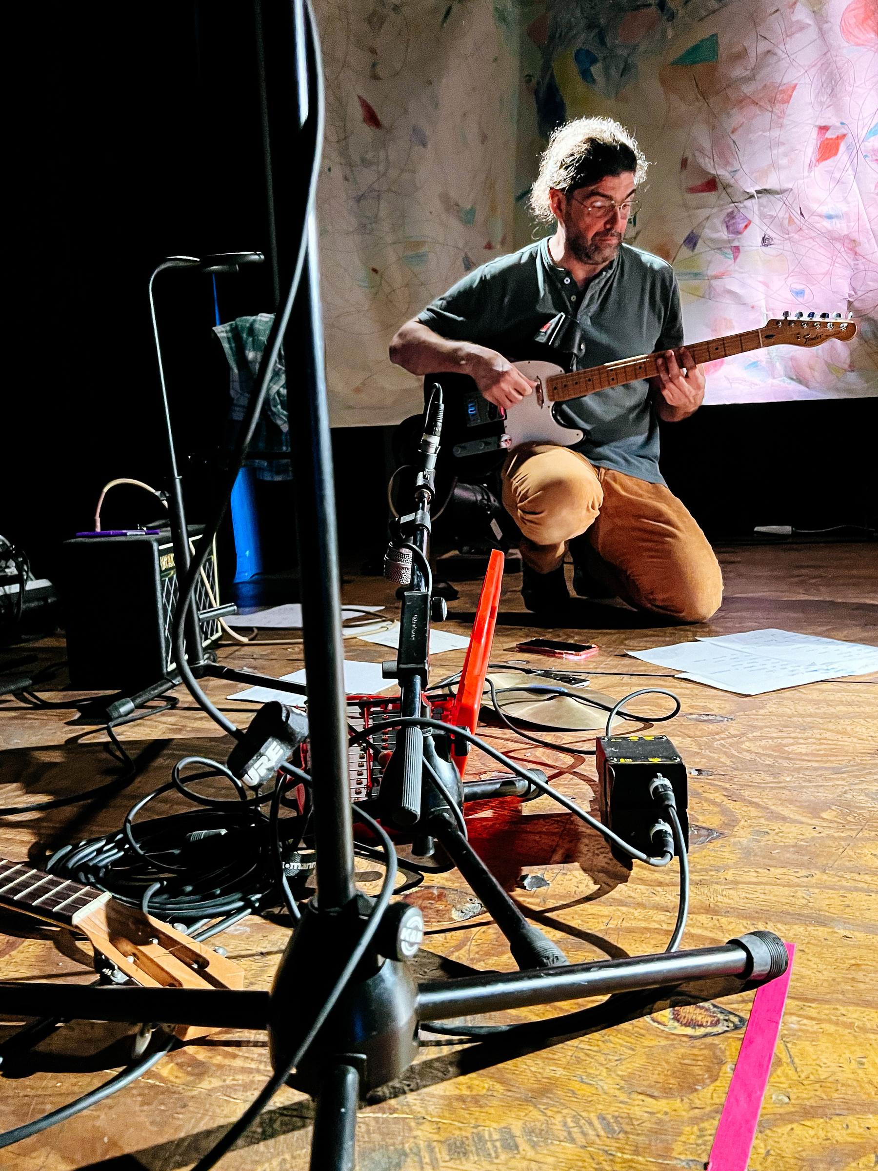 A guitar player kneeling down, plays his instrument, surrounded by amps and microphones. 