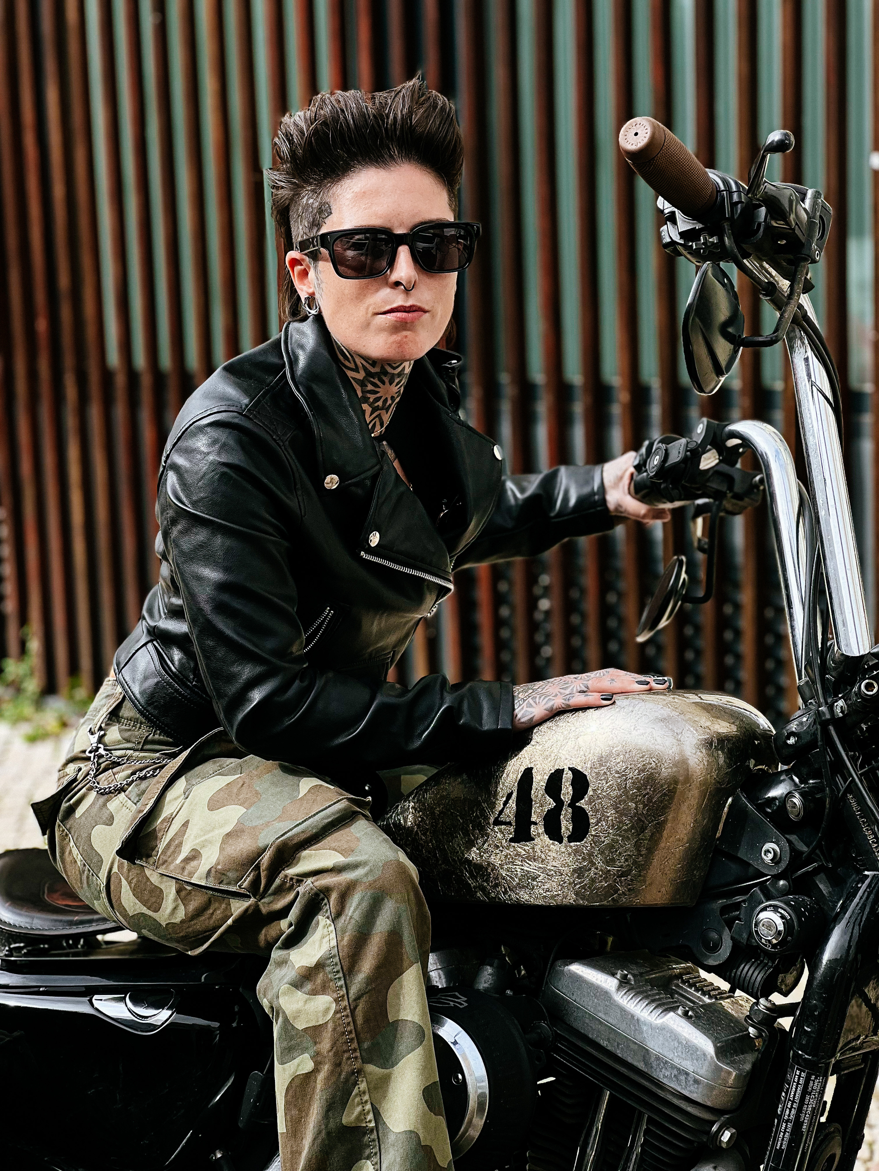 Girl wearing camo pants, leather jacket and shades sitting on a custom Harley, with 48 written on the tank 