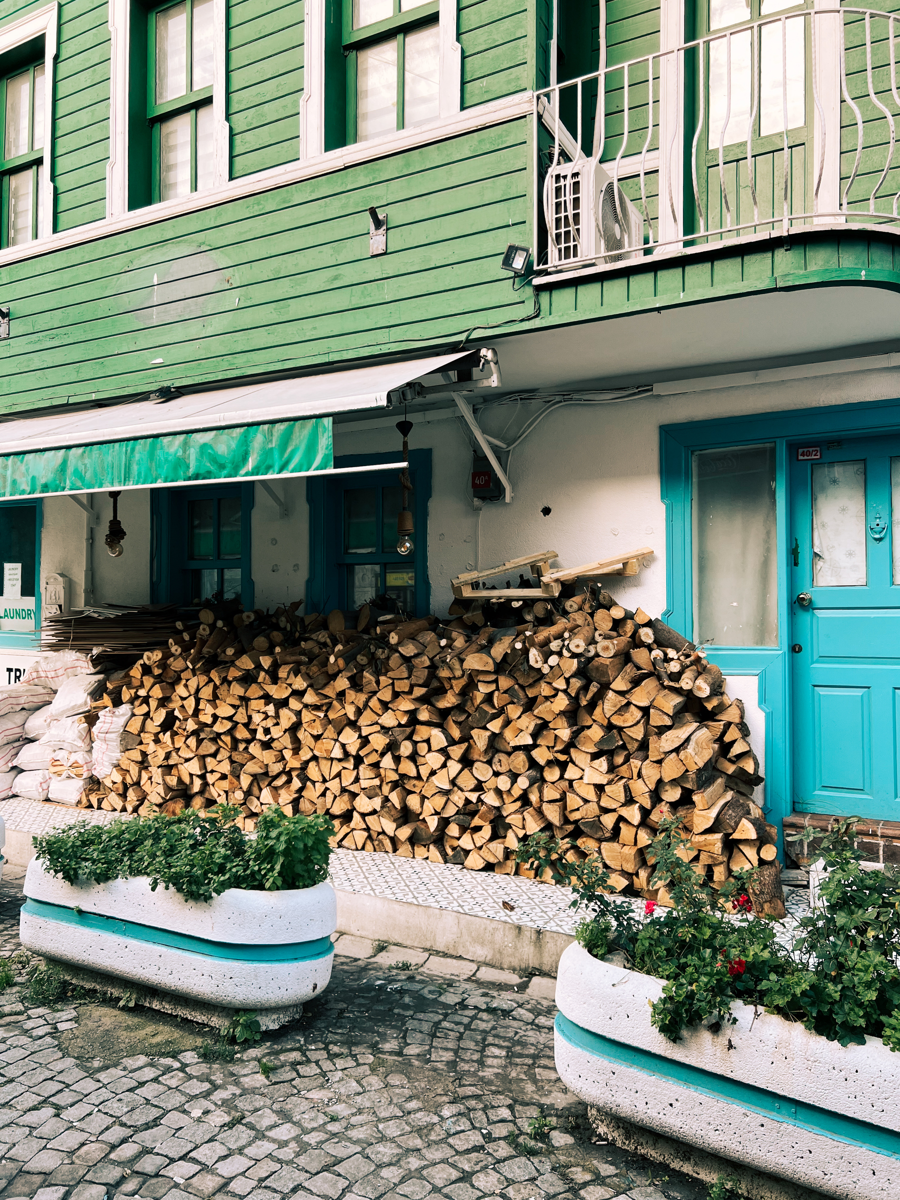 A stack of firewood piled up against the exterior wall of a building with green wooden cladding, beneath a balcony. Planters with greenery are situated in front of the woodpile.