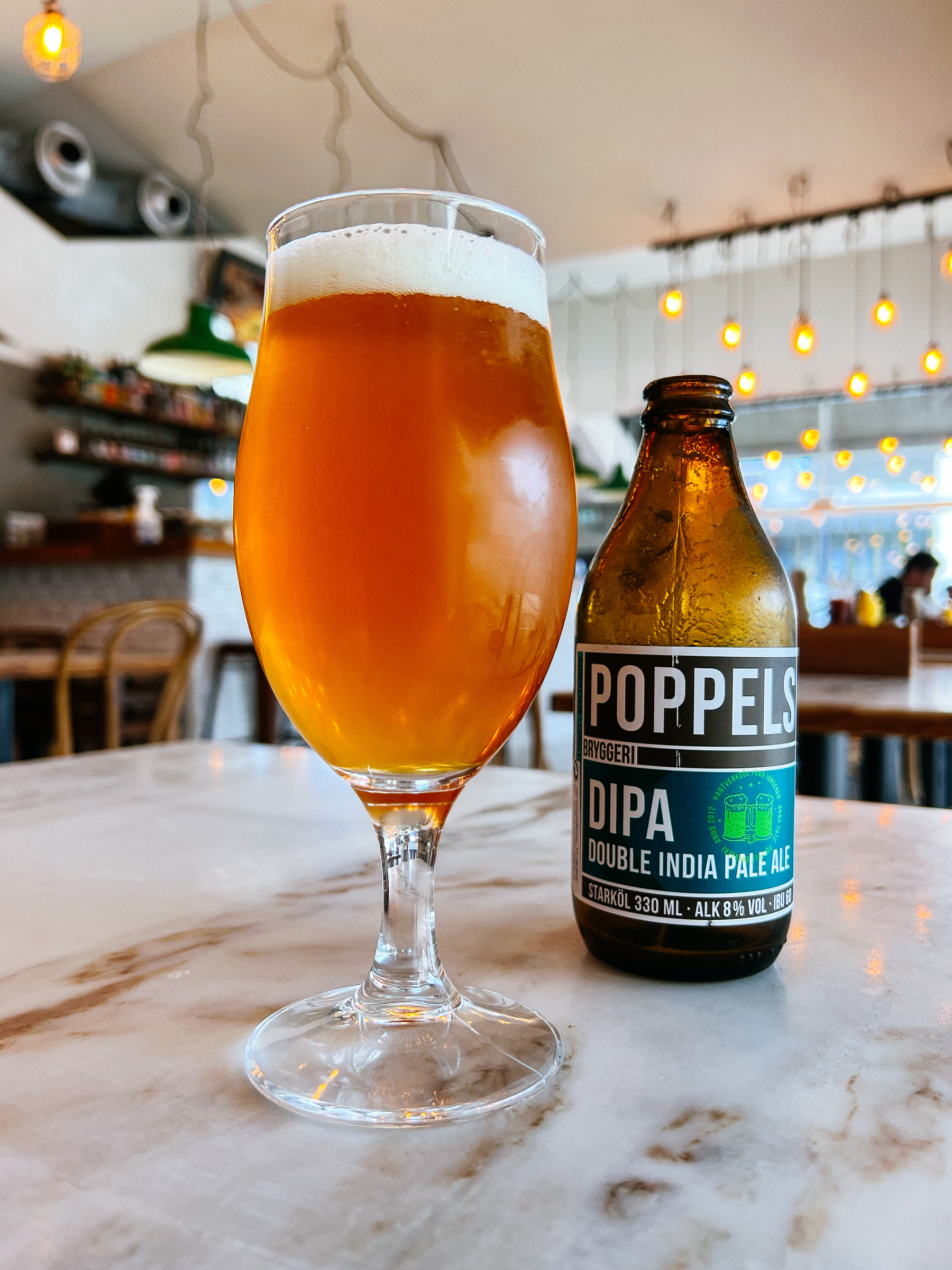 A beer glass, and a bottle by its side. Poppels, DIPA. Inside a restaurant. 
