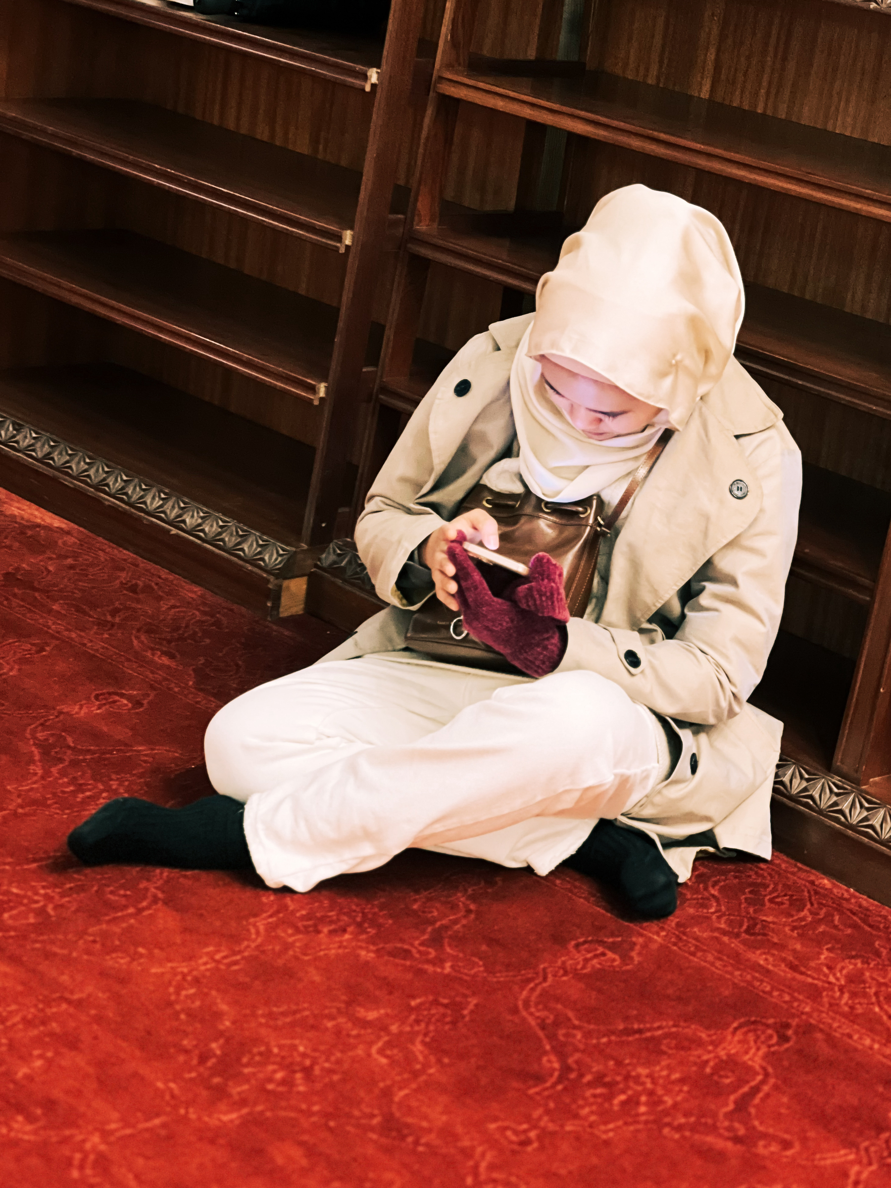 A person sits on a red carpeted floor against wooden shelves, checking their phone. The individual is wearing a beige trench coat, white pants, a white headscarf, and black socks.