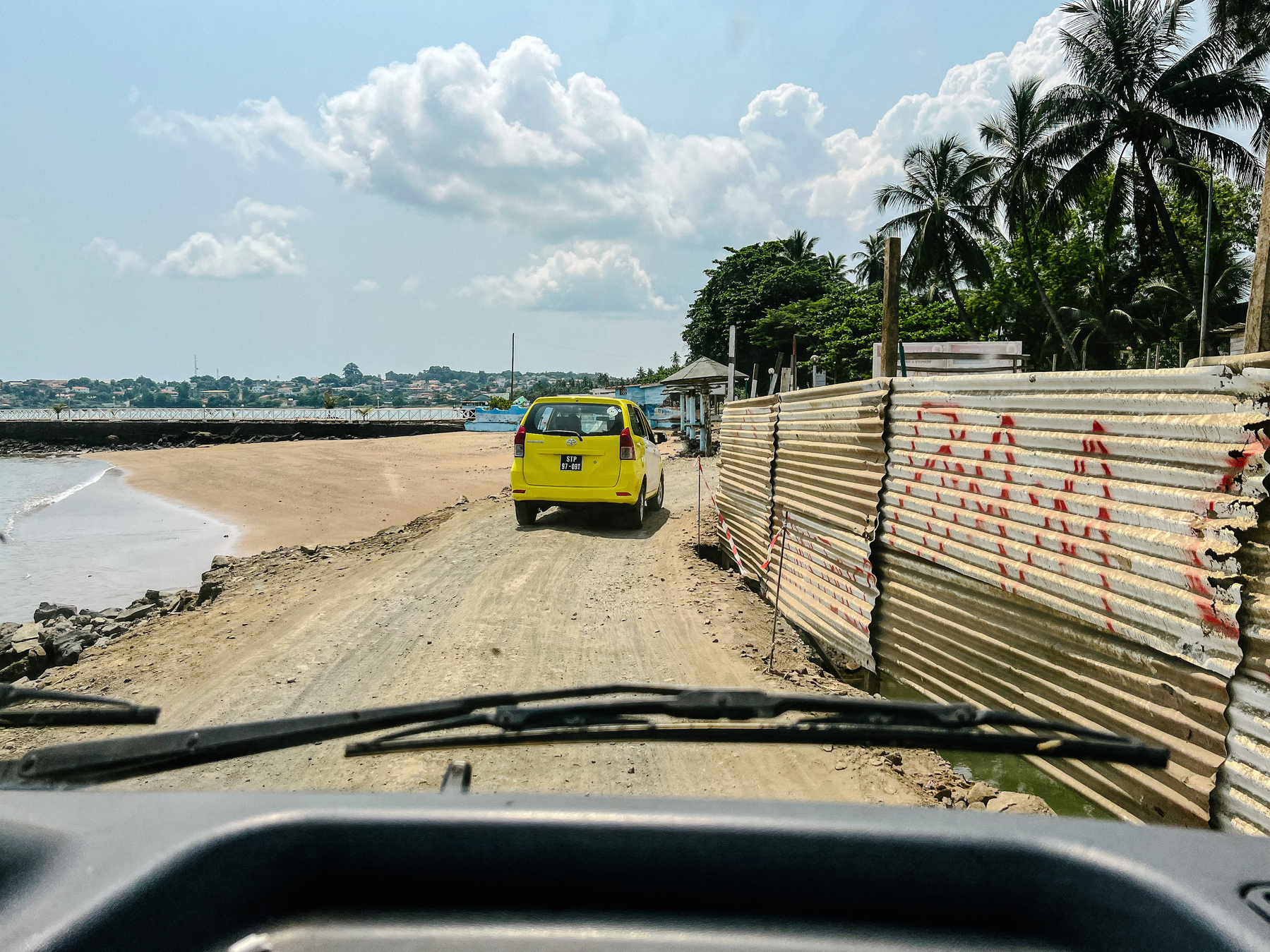 Seen from inside a car, we’re following a yellow car on a dirt road. The sea is on the left side. 