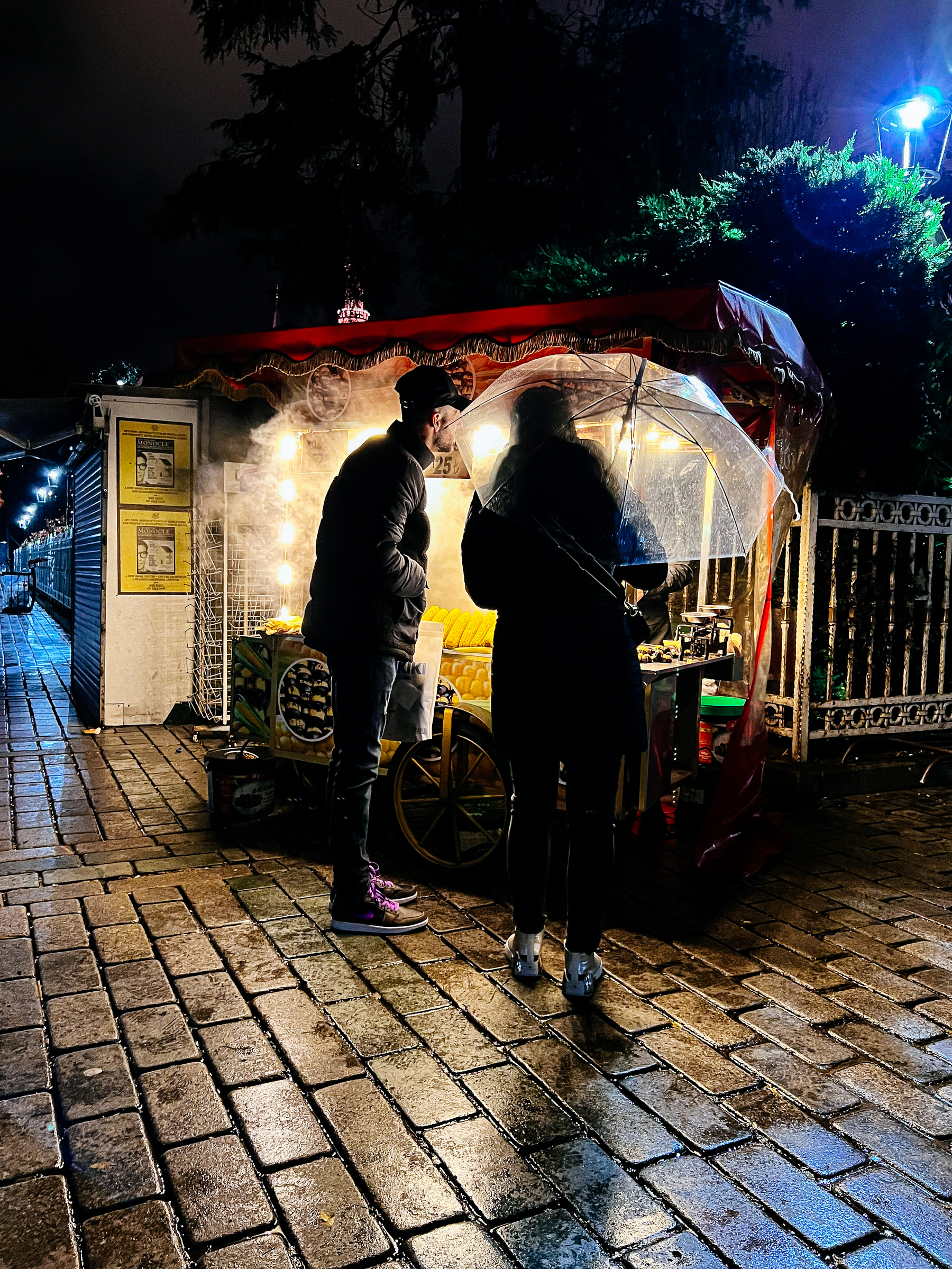 A couple by a roasted corn stall. The woman is holding an umbrella. 
