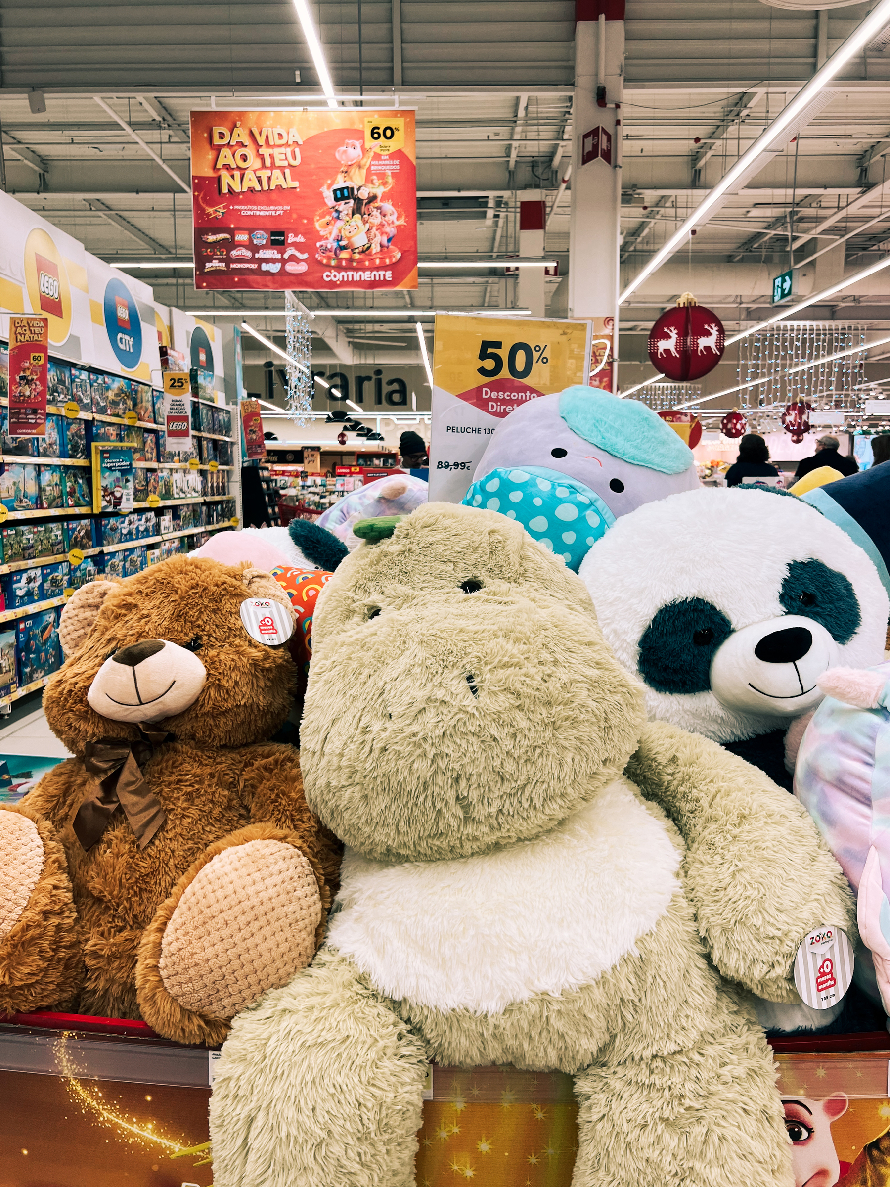 A selection of large plush teddy bears in a store, with a promotional sign advertising a 50% discount. The store appears to be decorated for a holiday season with banners and hanging decorations in the background.