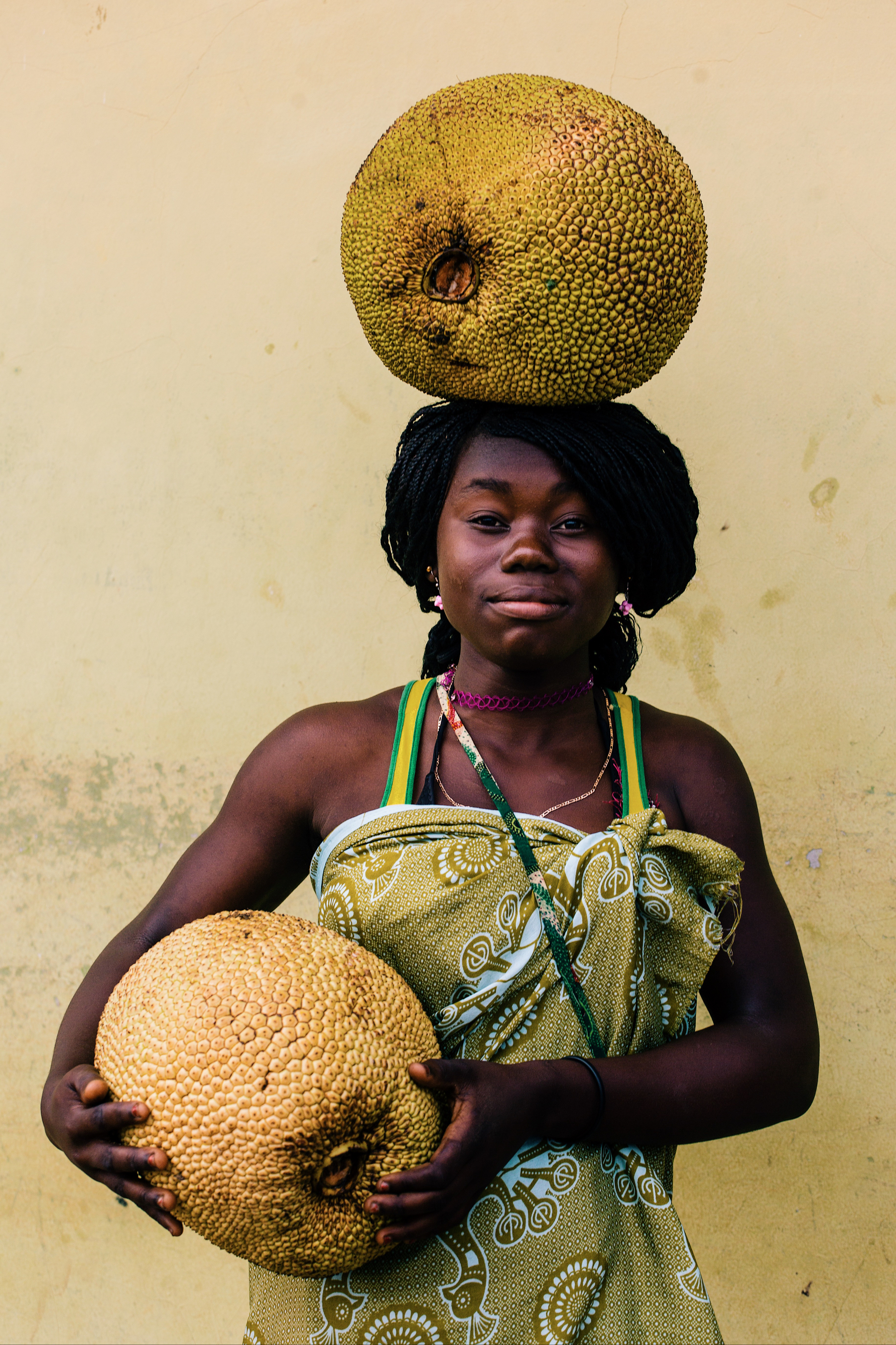 A woman carries a jackfruit on her head, and holds another one on her arms