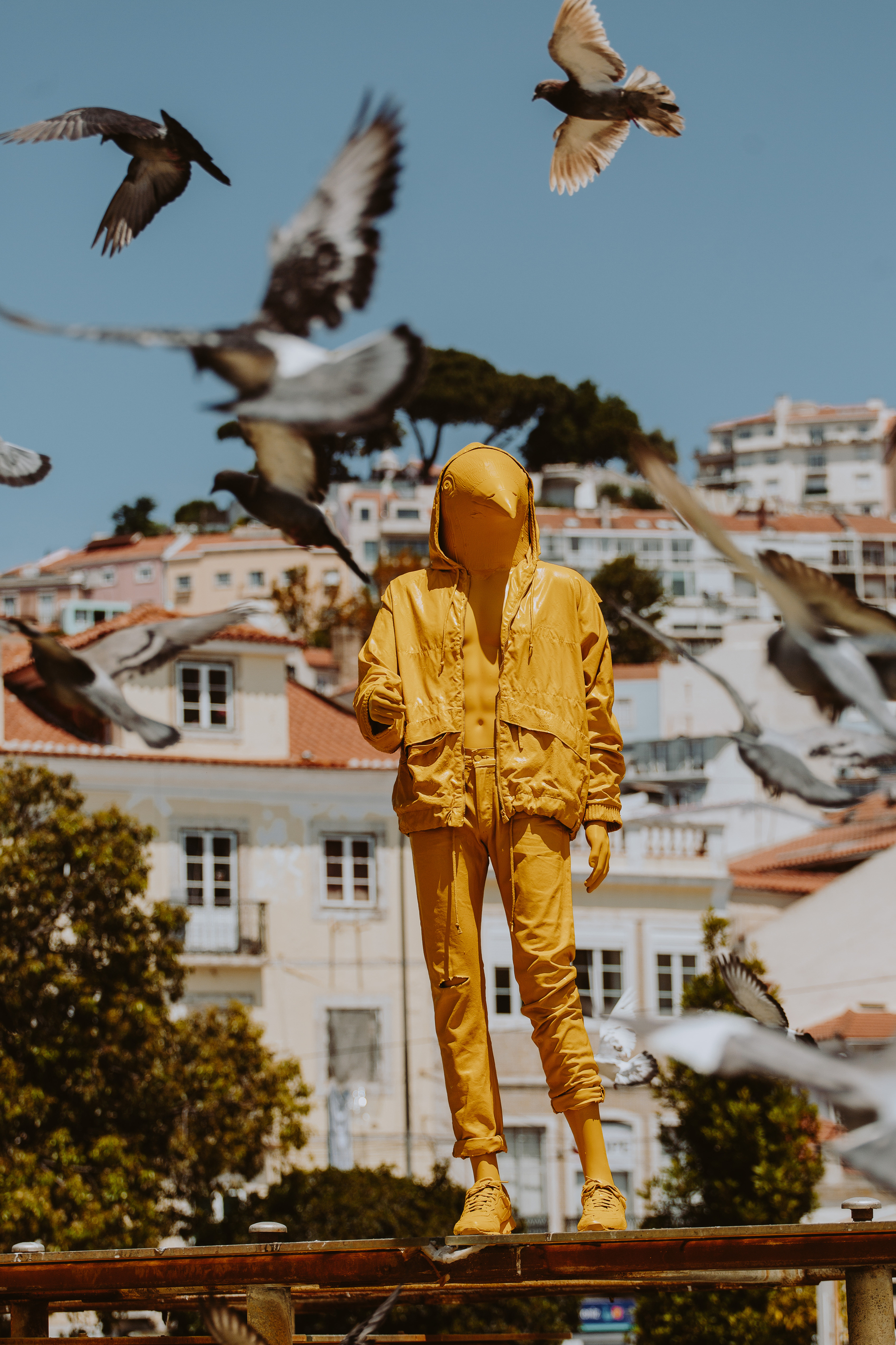 A street art installation: a yellow man, with a pigeon’s head. Cityscape in the background, and pigeons flying all around him. 