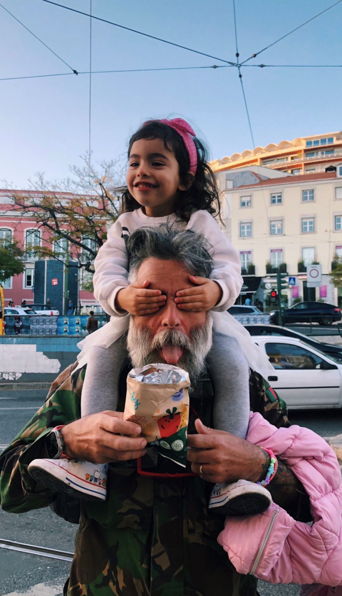 A toddler riding piggyback, covering her father’s eyes. 