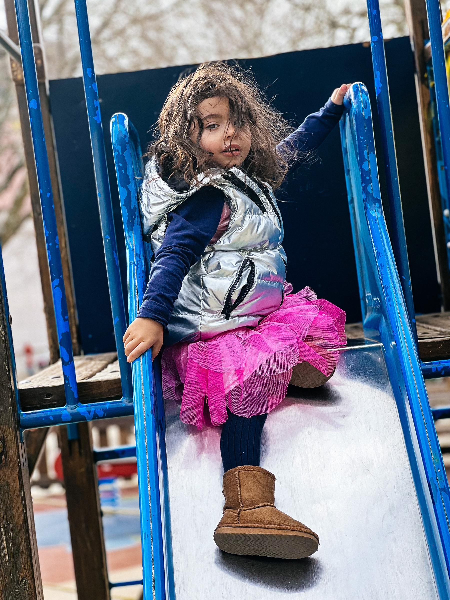 A girl sits at the top of a slide, looking at us. She’s wearing a pink skirt, and silver vest. 