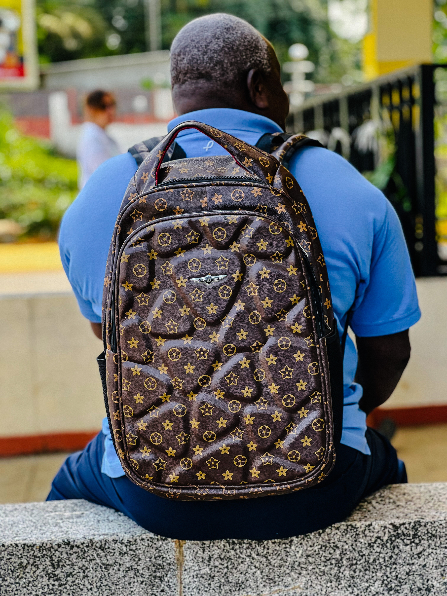 A man seen from the back, with an obviously very fake Louis Vuitton backpack. 