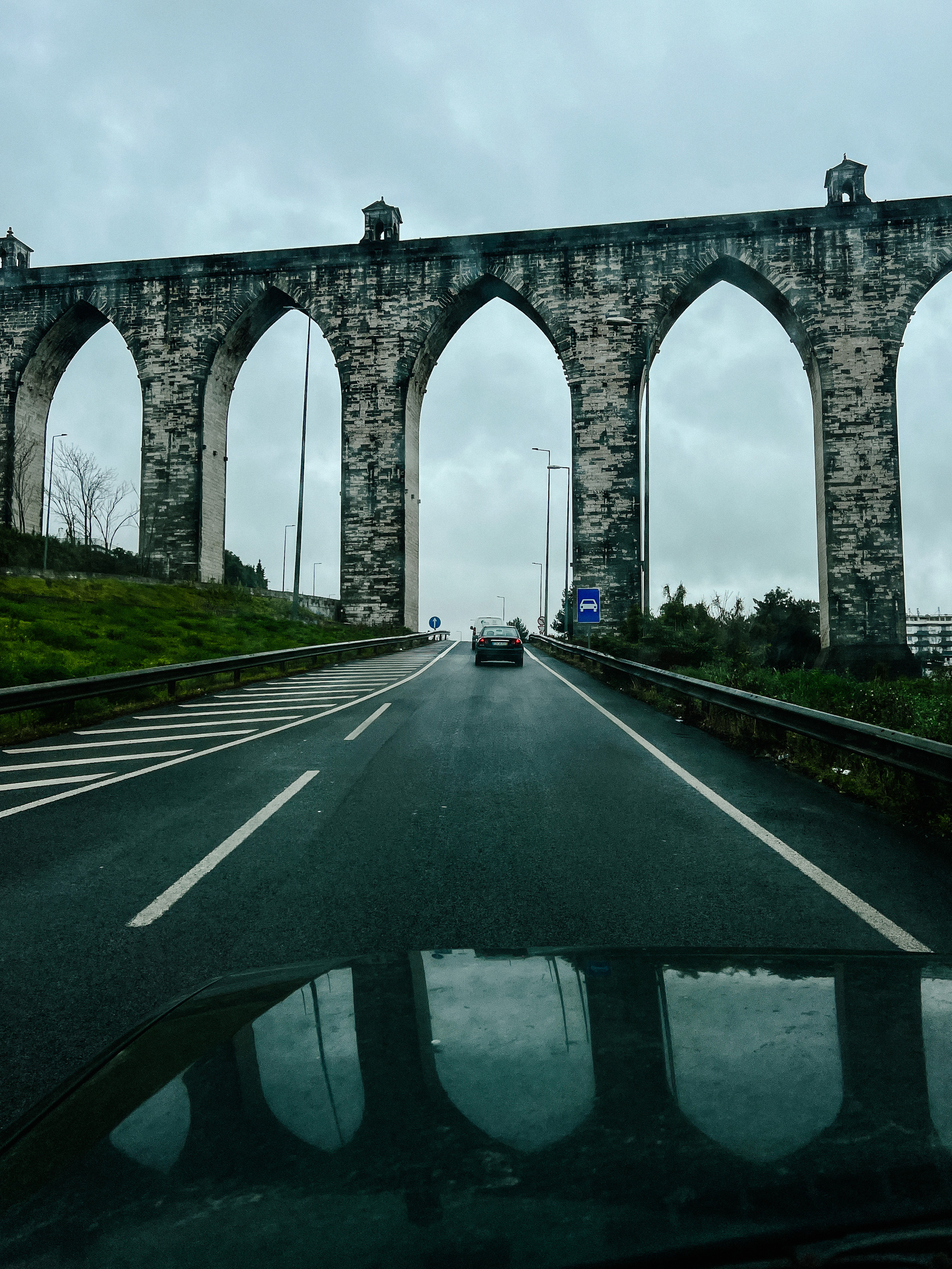 An aqueduct is seen ahead, while driving on a road. It’s reflected on the hood of the car from where the photo was taken. 
