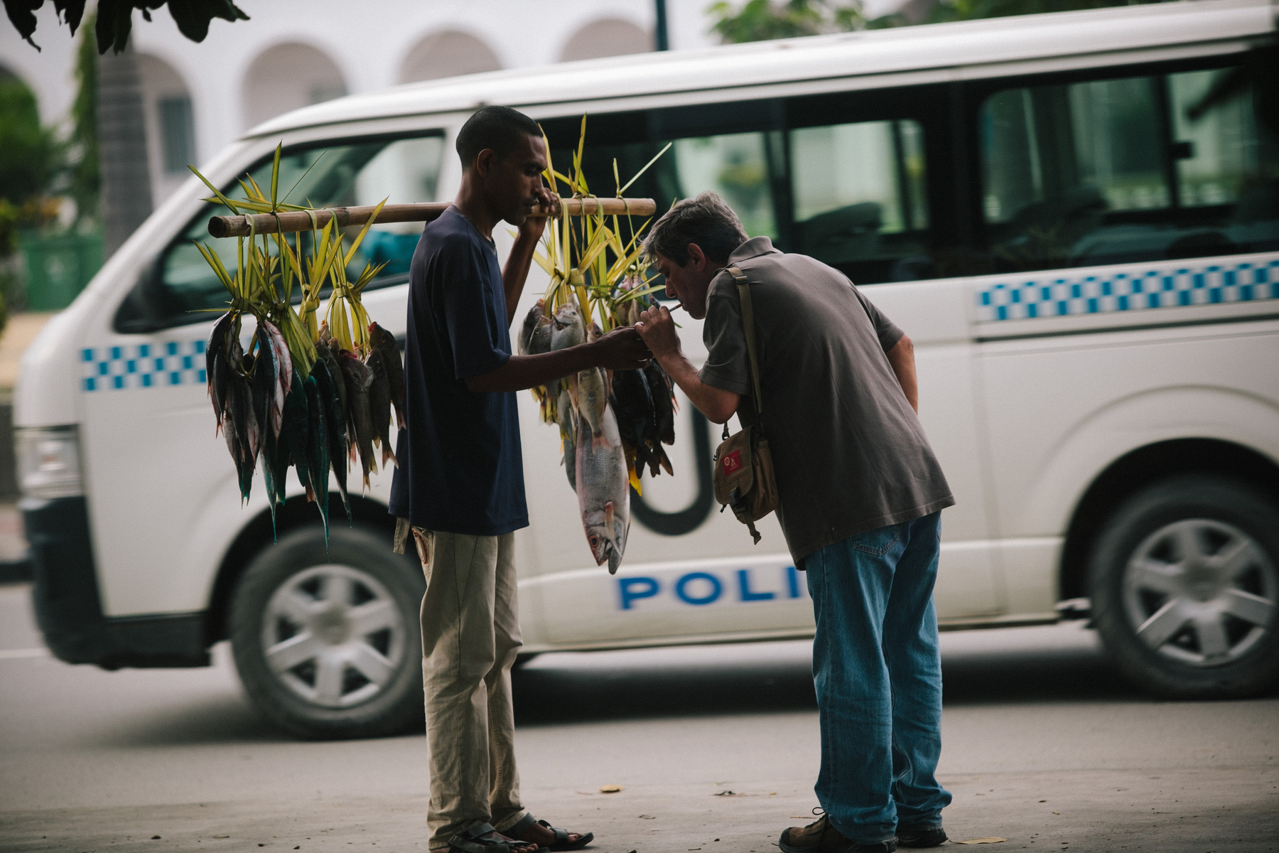 A man selling fish lights up a cigarette belonging to another man. A police van drives by. 