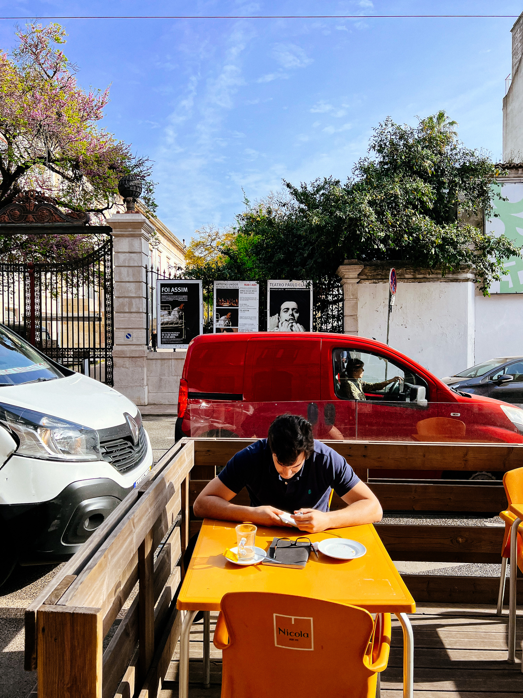 A man writes on a notebook, sitting outside at a cafe. Cars going by in the back. 