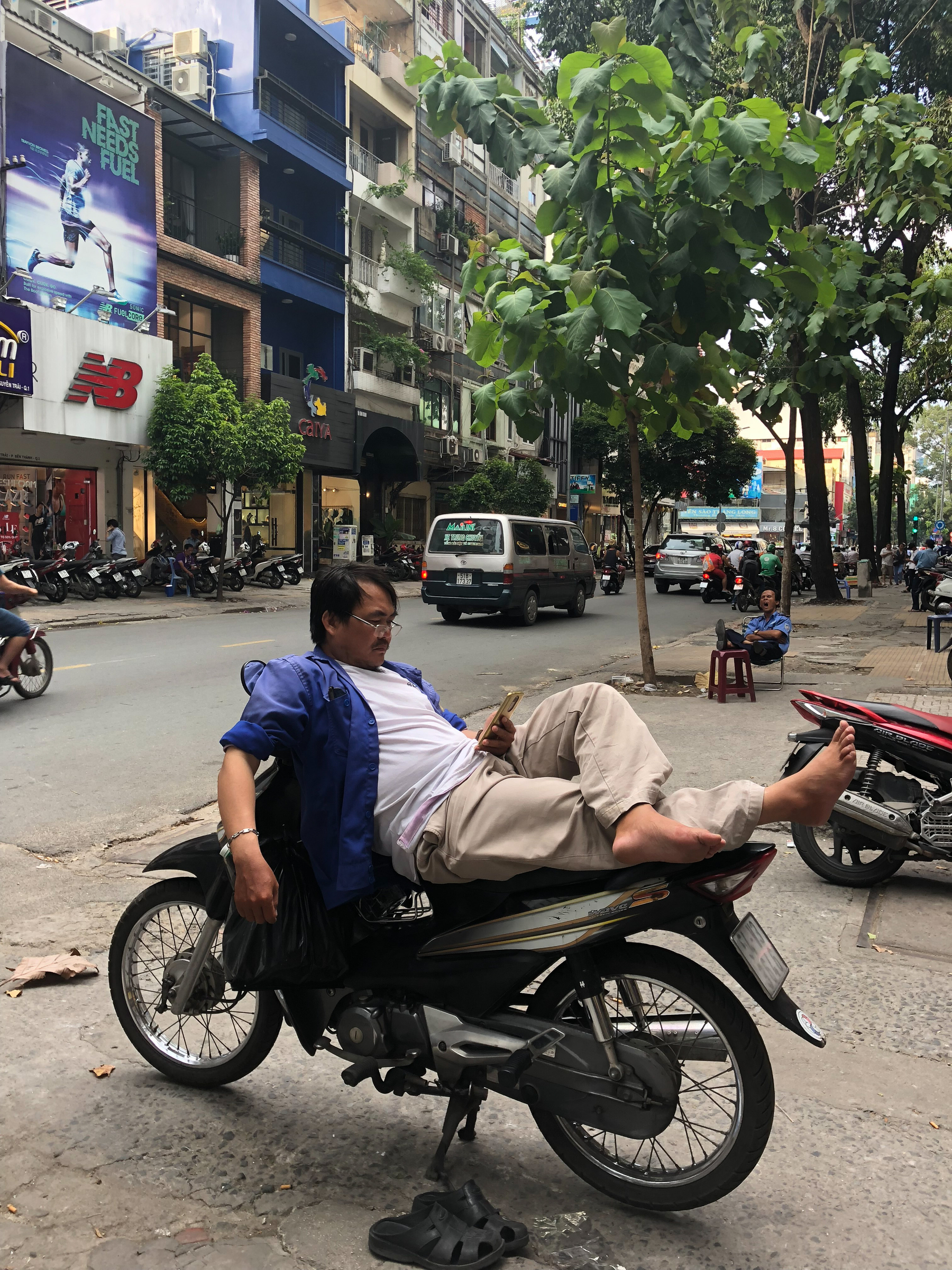 A man checks his phone while lying down on top of his motorcycle, in a busy city. 