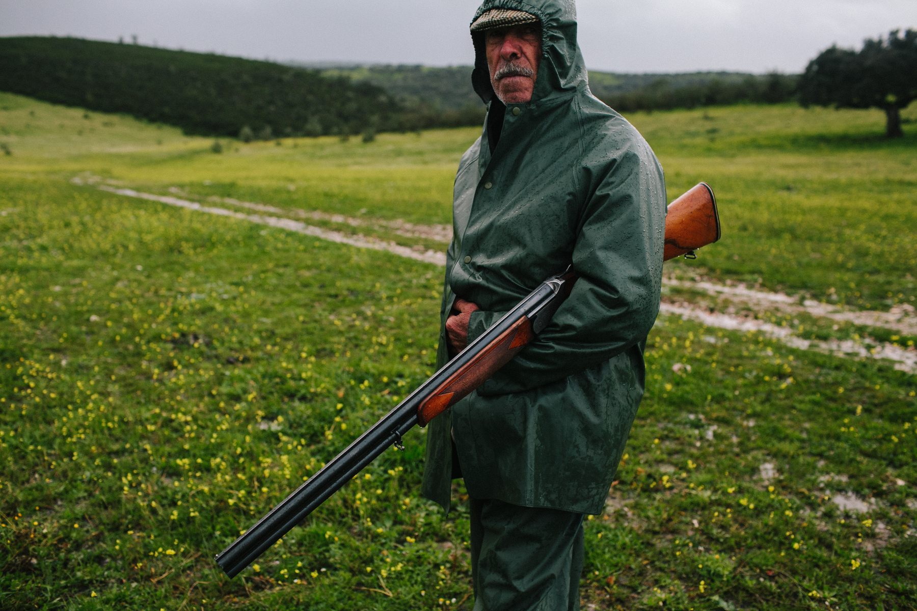 a man holds a shotgun in the middle of a field. He’s wearing a green raincoat, and the whole landscape is also green