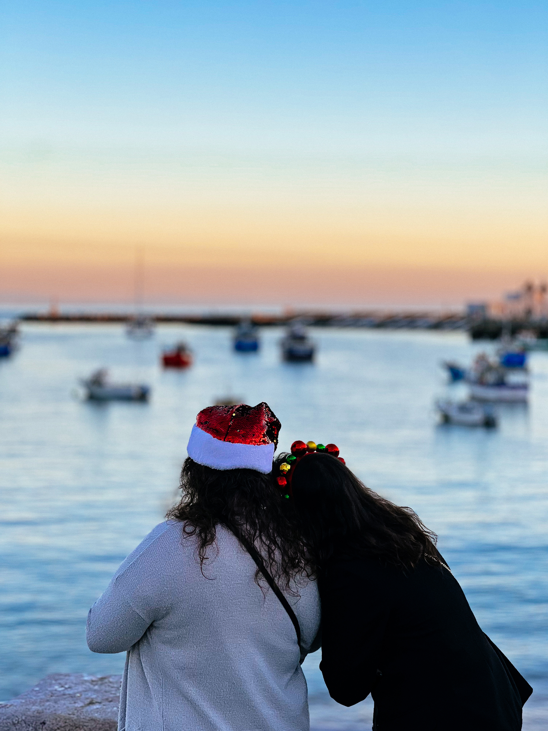 Two individuals wearing festive holiday headwear looking out at a harbor with boats during sunset.