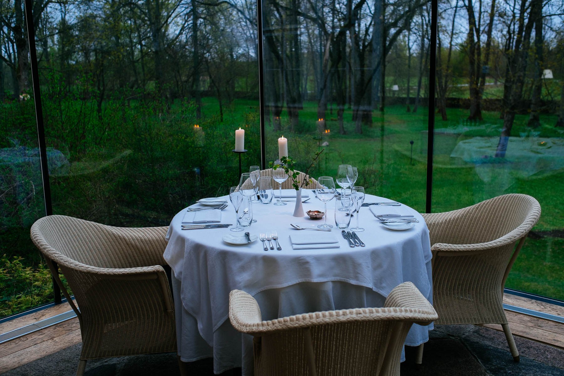 a table is set on a restaurant, with a huge glass window next to it, and a glorious view to the forest outside