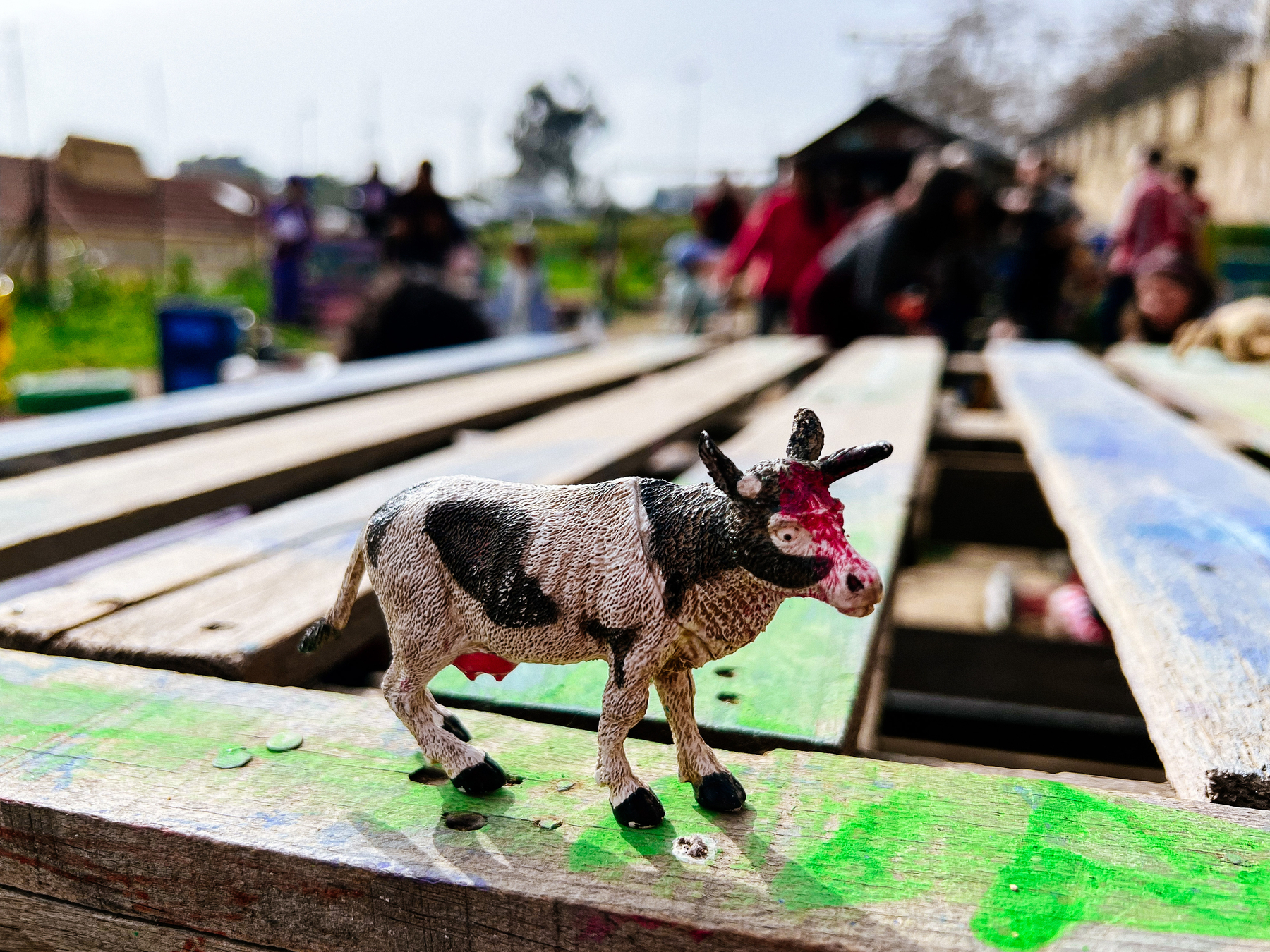 A toy cow stands in a wooden structure. People out of focus in the back. 