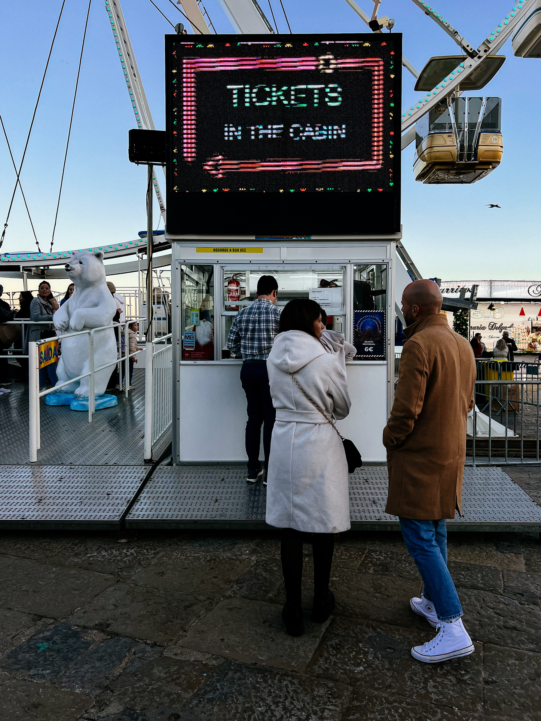 Queue of people at a ticket booth for a Ferris wheel under a bright LED sign that reads &ldquo;TICKETS,&rdquo; with a large Ferris wheel cabin visible in the background and a polar bear statue to the left.