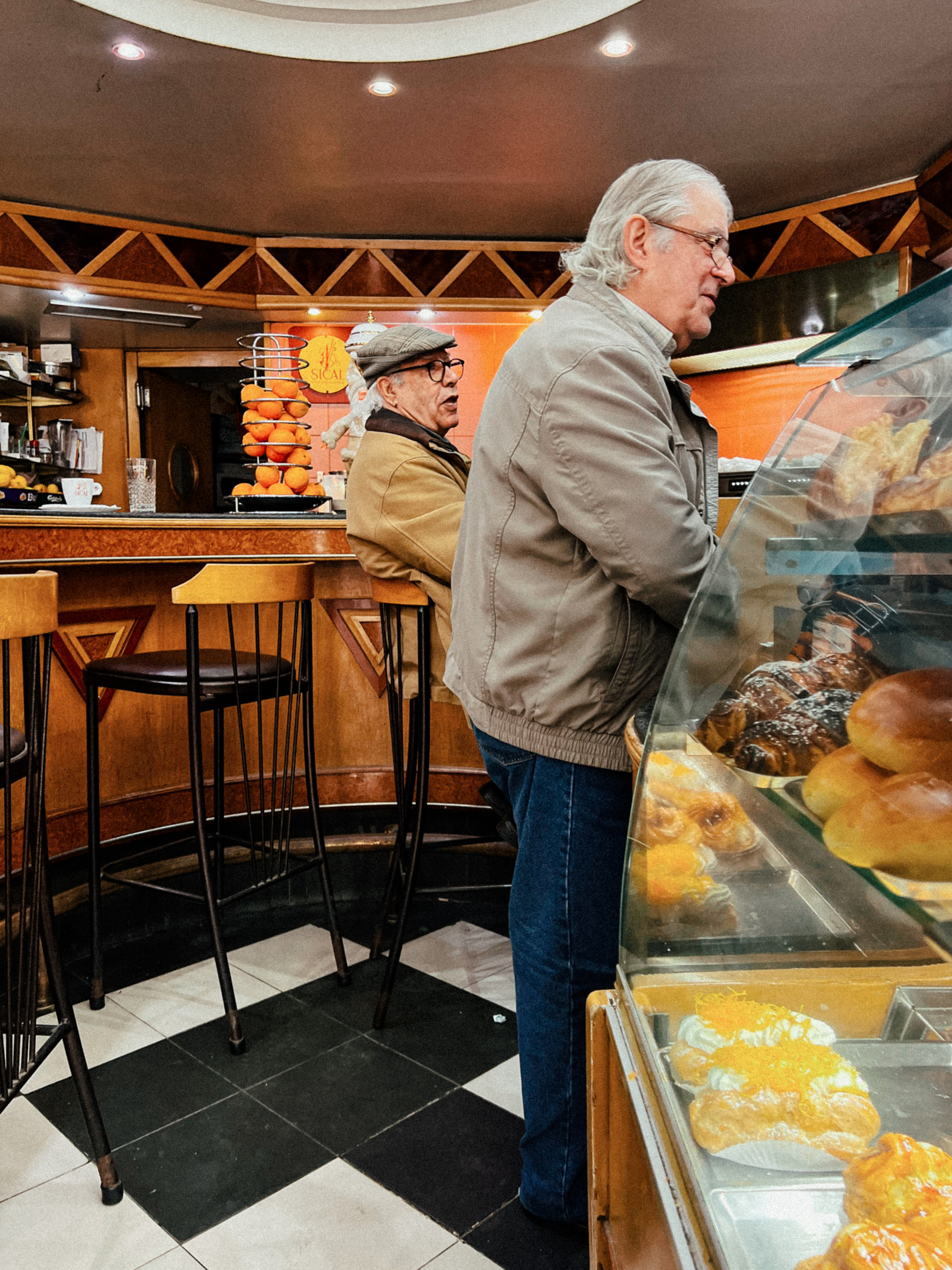 Two men stand at the counter in a classic, wooden, coffee shop. Cakes in the foreground. 