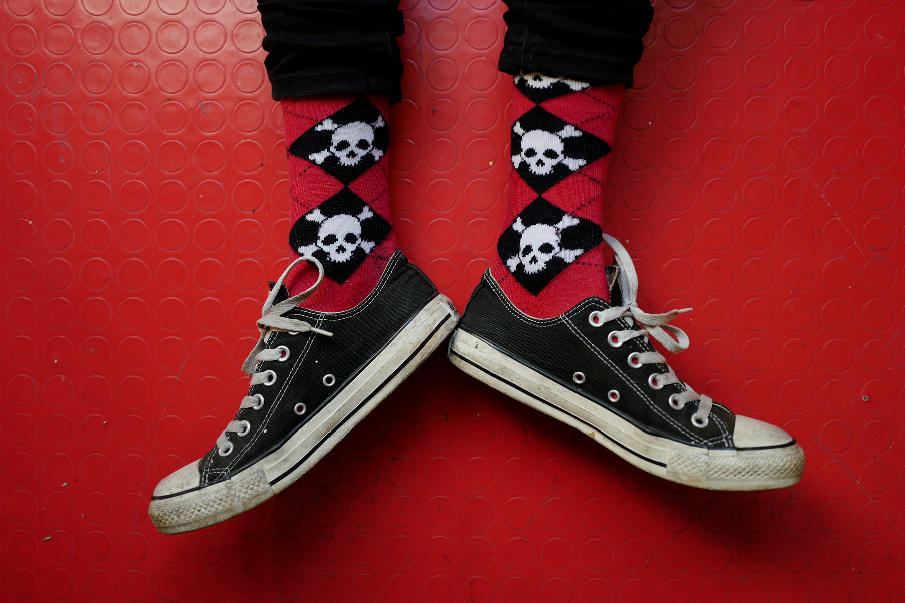 Looking down on a pair of legs with sneakers, red floor, red socks with skulls.