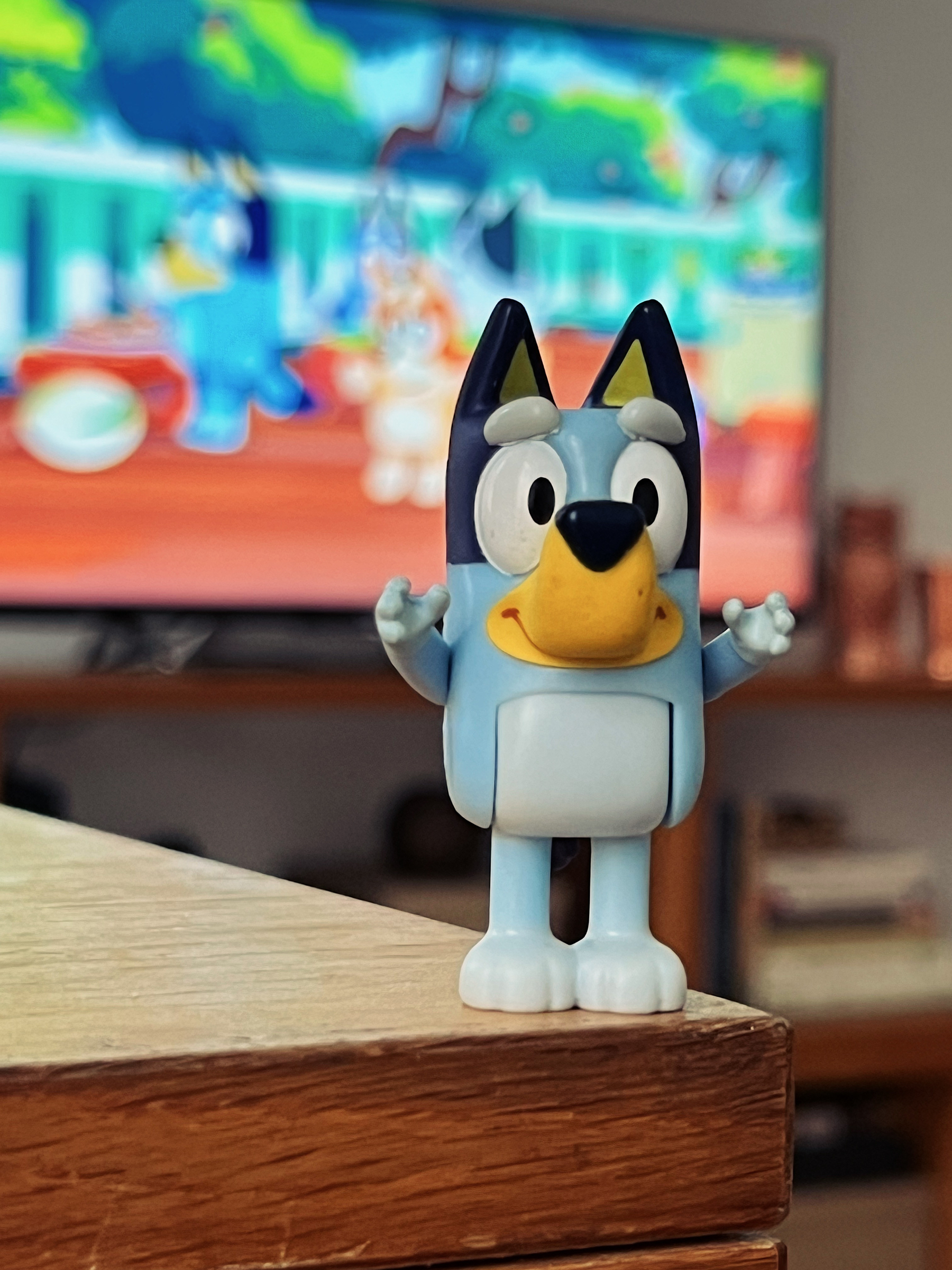 Bluey stands on a table, while Bluey plays in a tv in the background. 