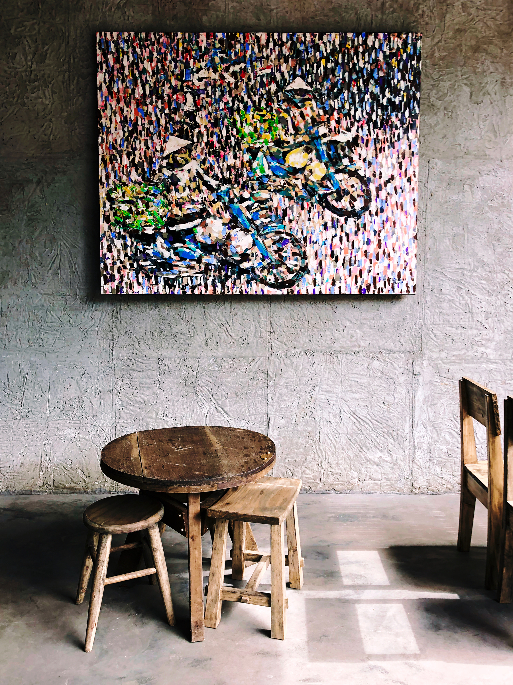 A wooden table, and two wooden stools, sit in front of a painting. 
