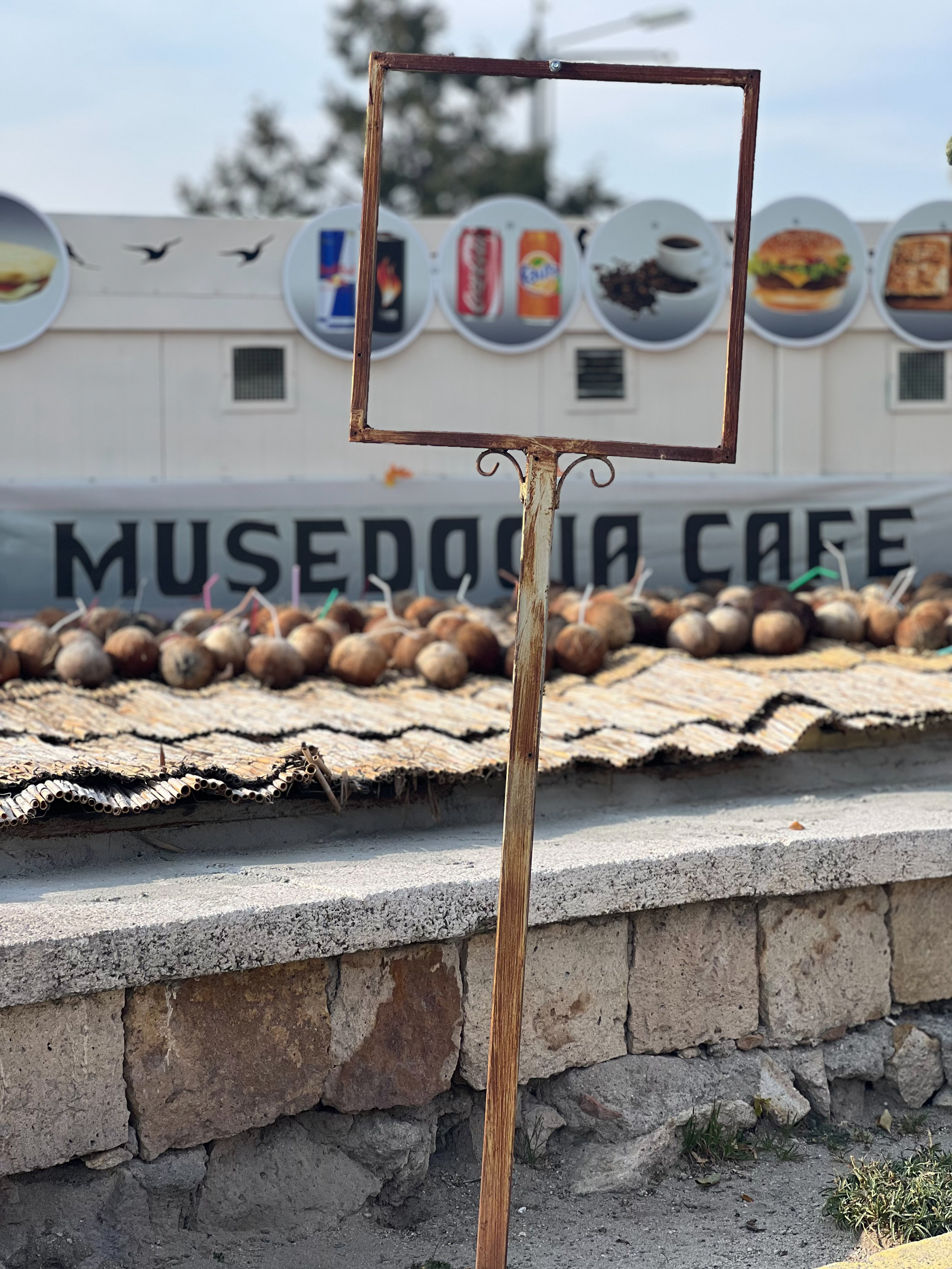 A rusty square metal frame on a stand in front of a café, with a backdrop of advertising dishes and beverages. The café&rsquo;s name, &ldquo;MUSEDOCIA CAFÉ,&rdquo; is partially visible in the background, and coconuts.
