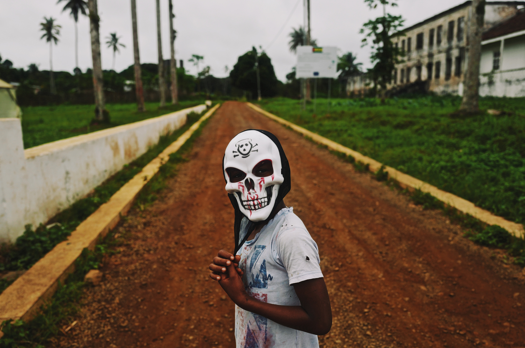 A kid wearing a skull mask, with a dirt road behind him. 