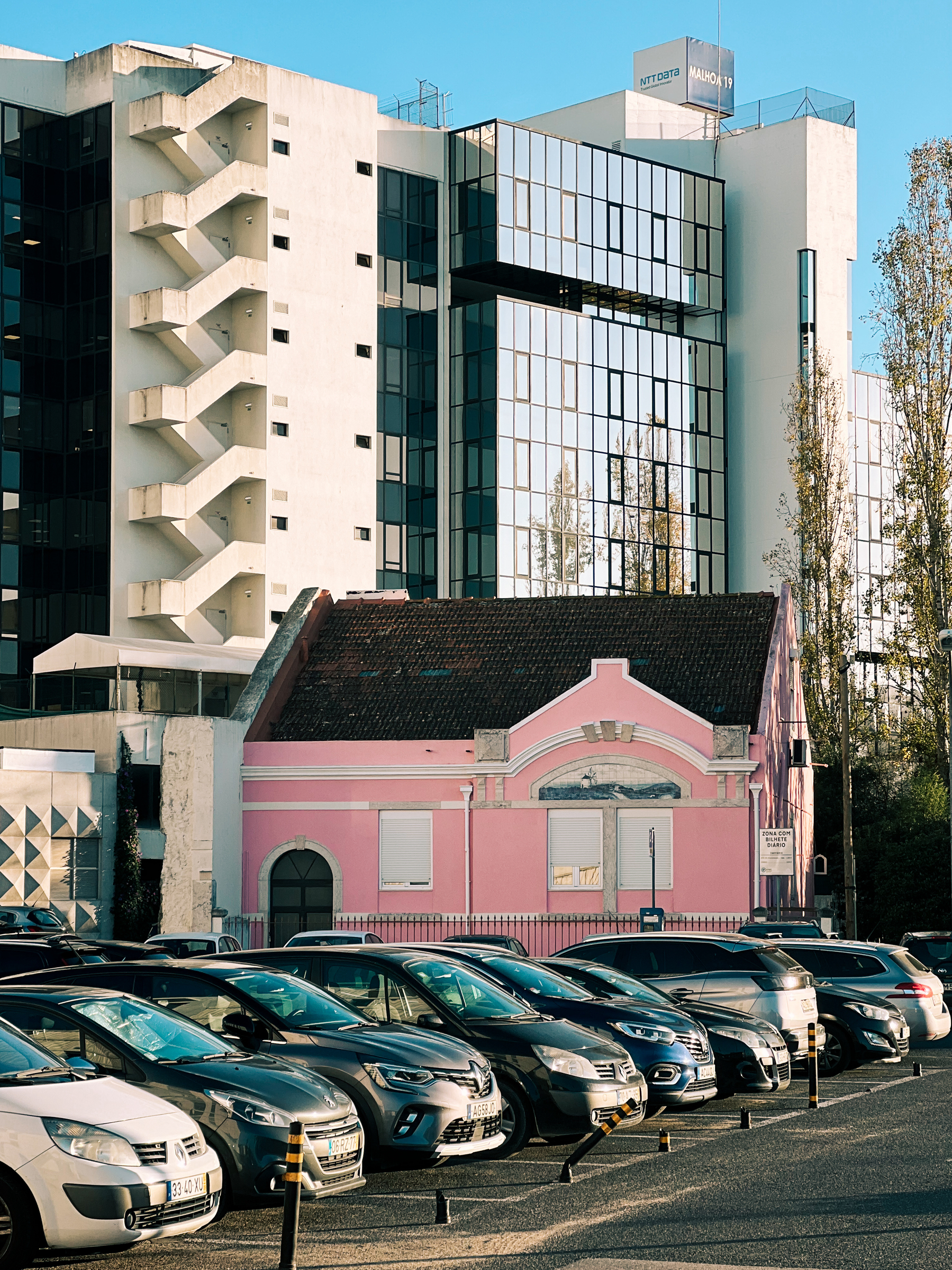 An old pink house stands in the middle of a parking lot, surrounded by modern looking buildings. 