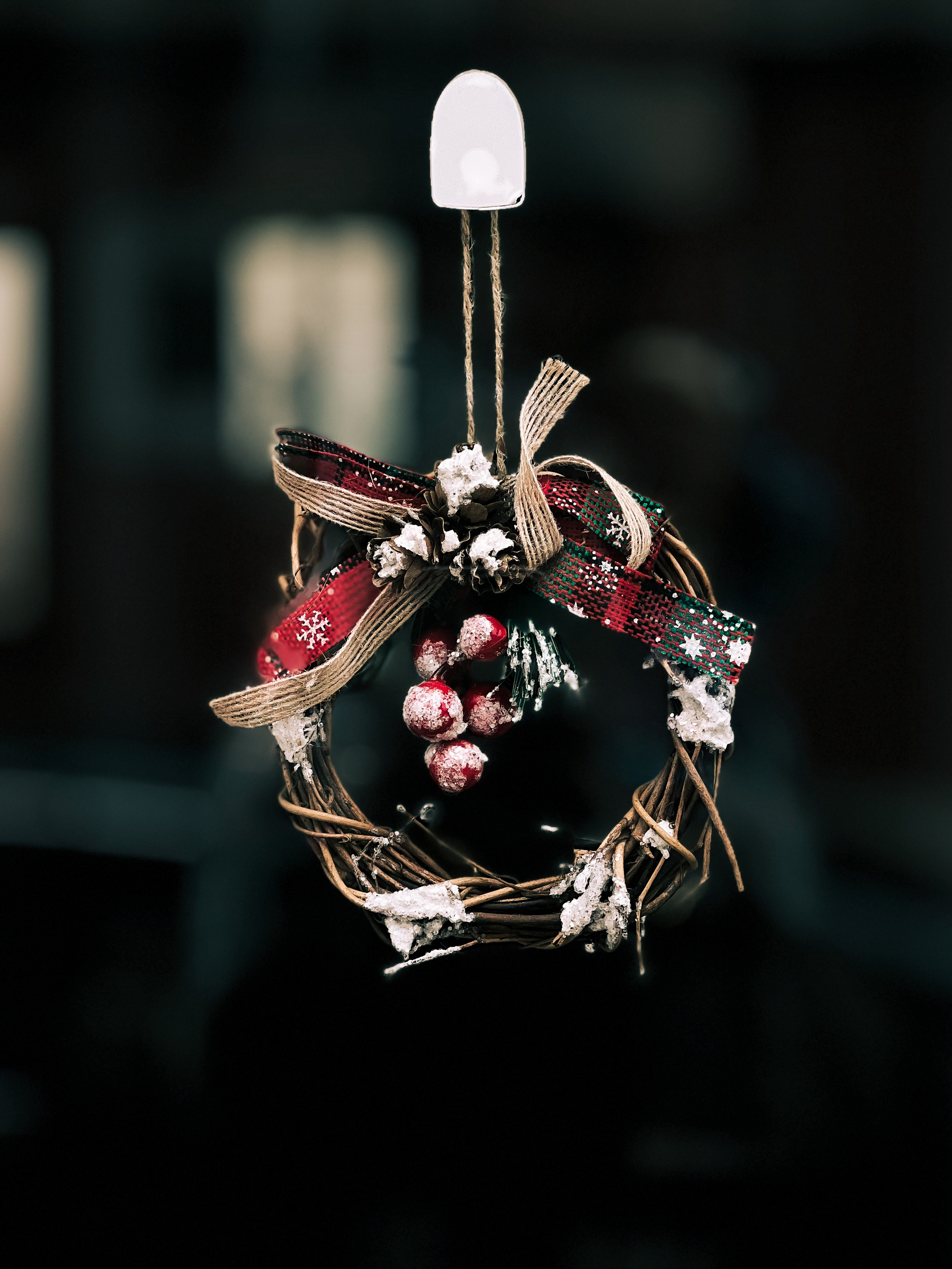 A festive Christmas wreath with red and tartan ribbons, frosted pine cones, and red berries, hanging by a twine loop with a blurred background.
