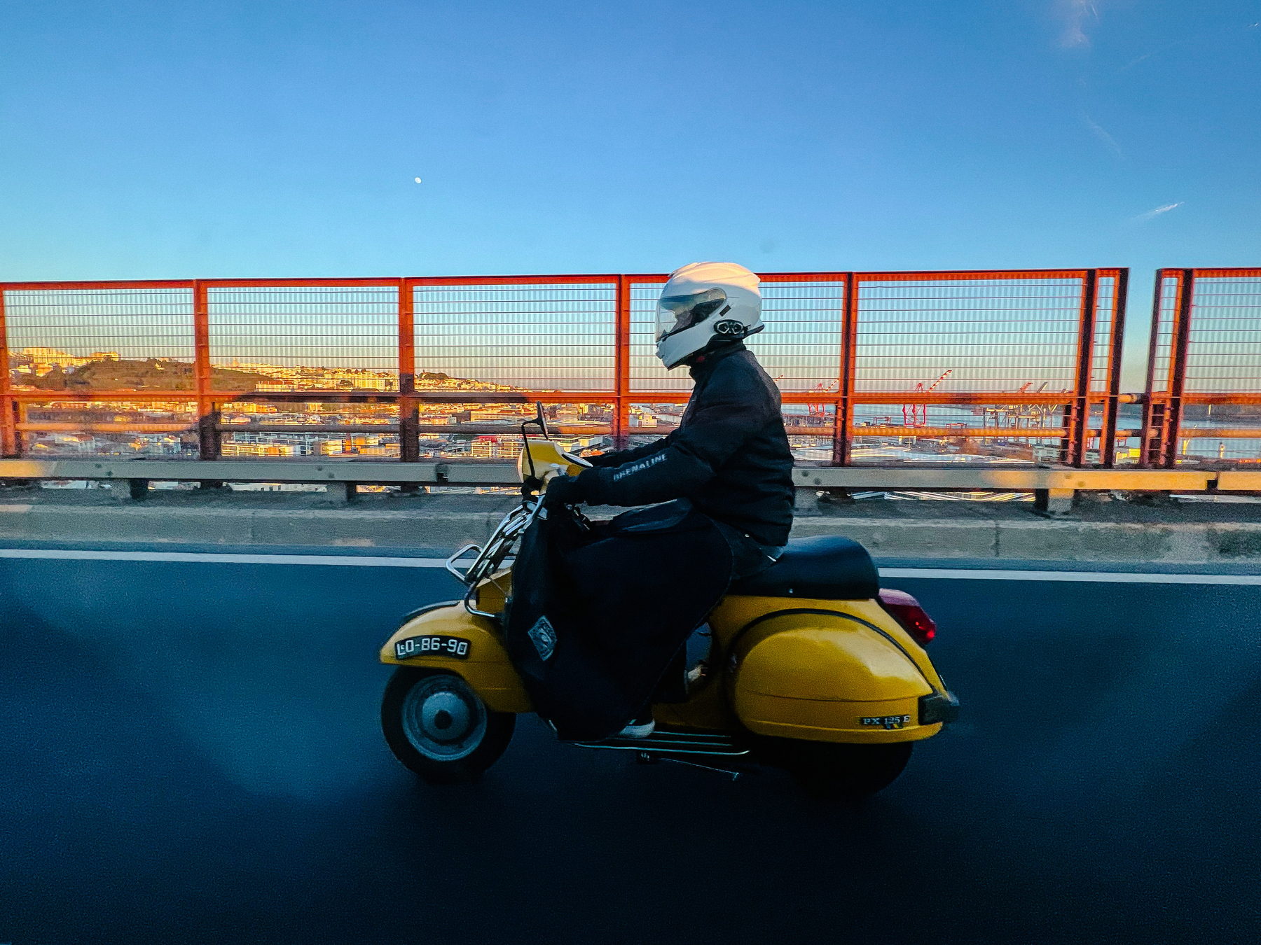 A yellow Vespa riding on a bridge, the city visible in the distance. 