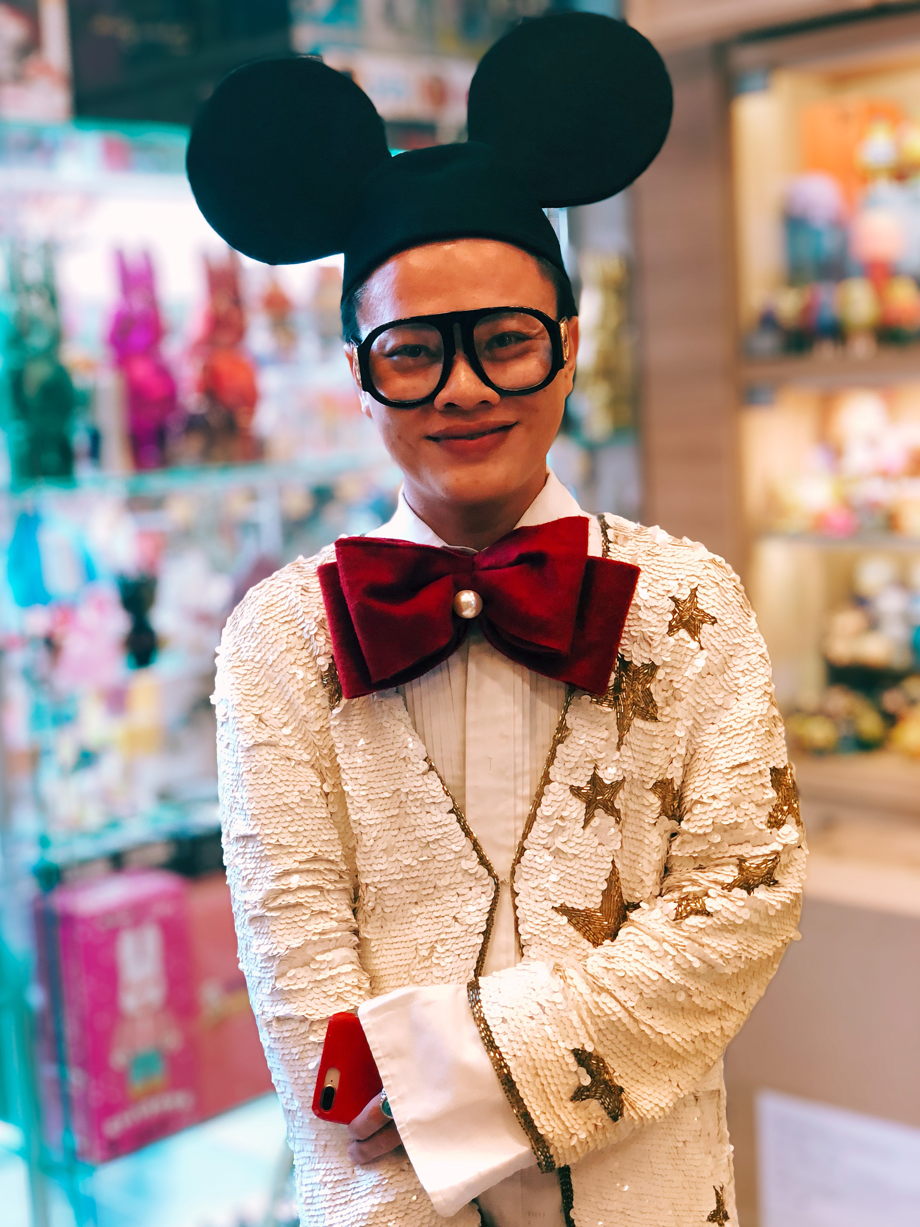 A young man dressed in a white jacket, with an oversized red bow tie, and Mickey Mouse ears. Big glasses too. 