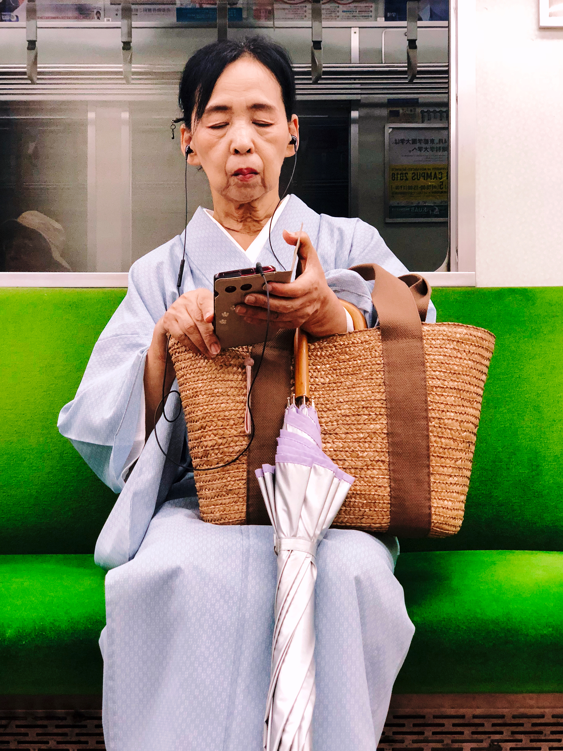 A woman in traditional Japanese outfit sits on a green train seat, holding a bag, an umbrella, and her phone. She has headphones on, and her eyes are closed. 
