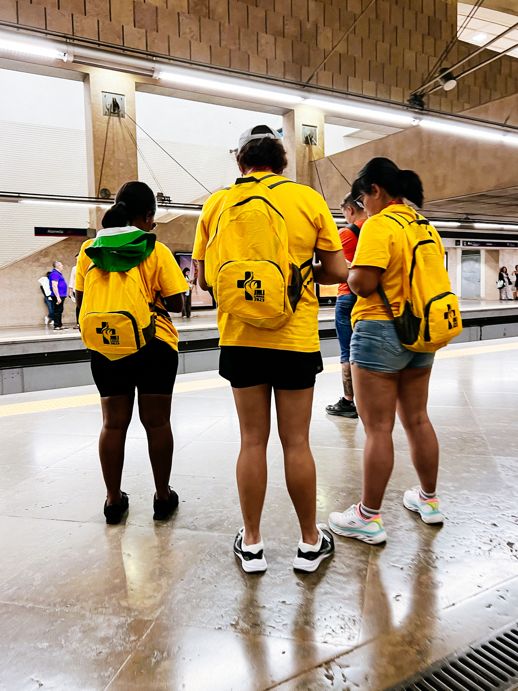 Three people wearing yellow t-shirts and yellow backpacks, waiting for the subway. 