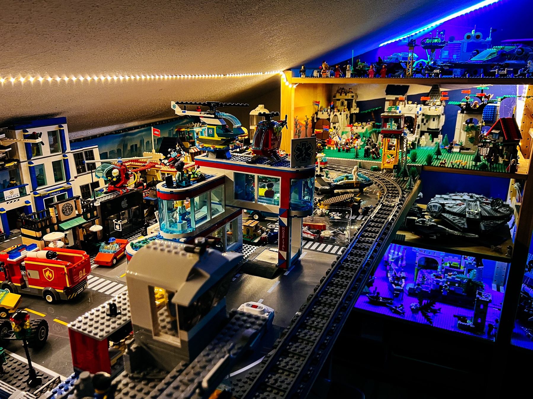 A detailed LEGO display featuring an urban cityscape with buildings, a fire station, vehicles, rail tracks, and a backdrop of a Star Wars Millennium Falcon model and a festive holiday scene. Bright LED lights illuminate the setup.