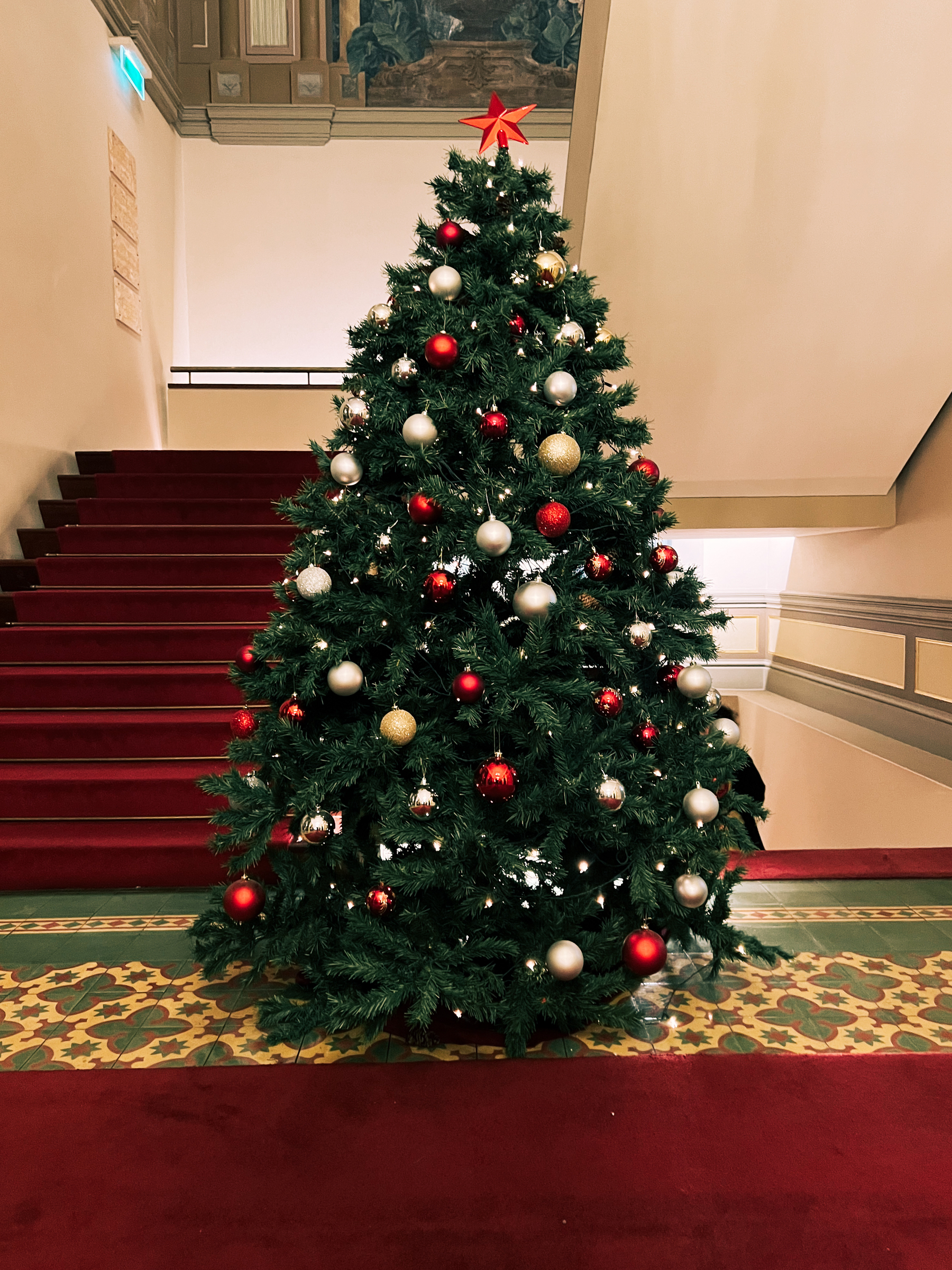 A Christmas tree next to a red carpeted flight of stairs. 