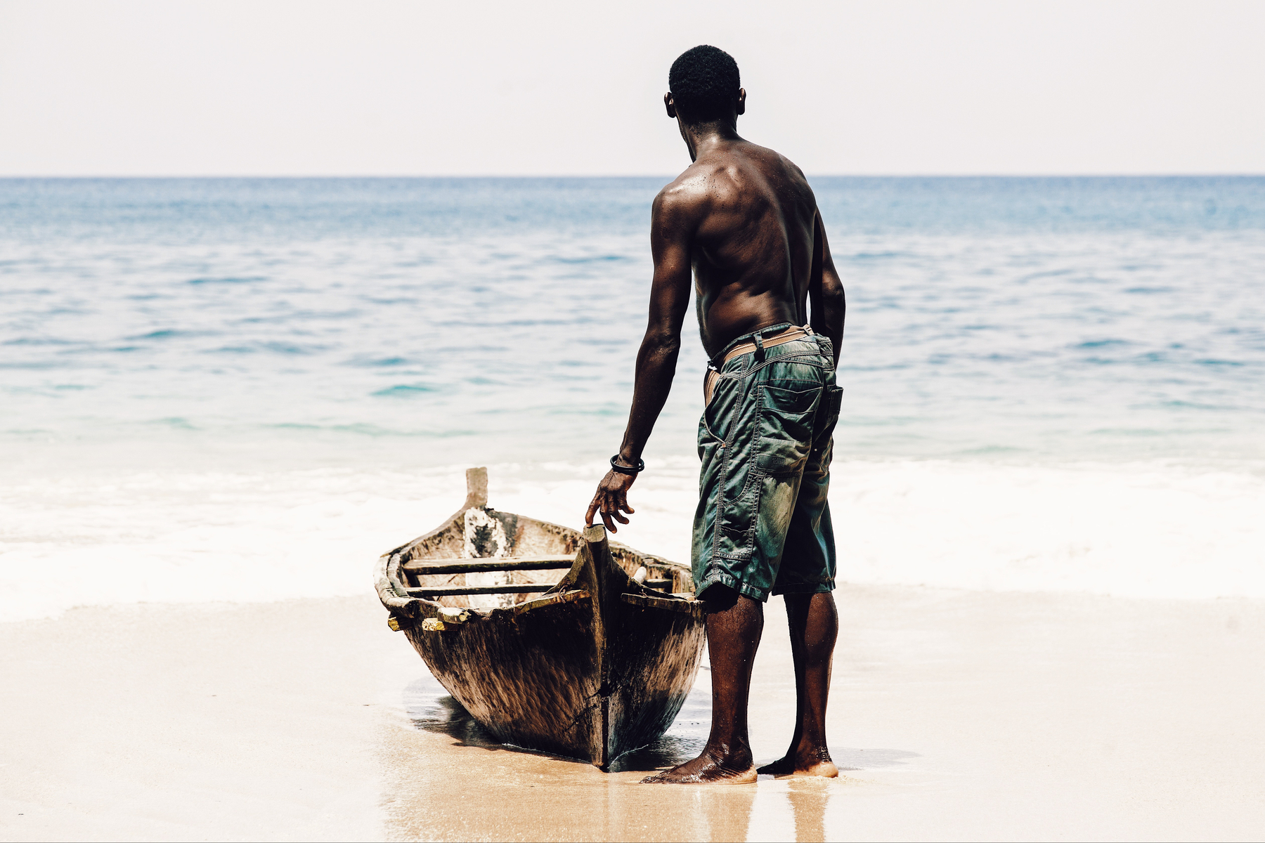 A man stands next to his canoe, gently touching the boat. He’s looking out into the sea. 