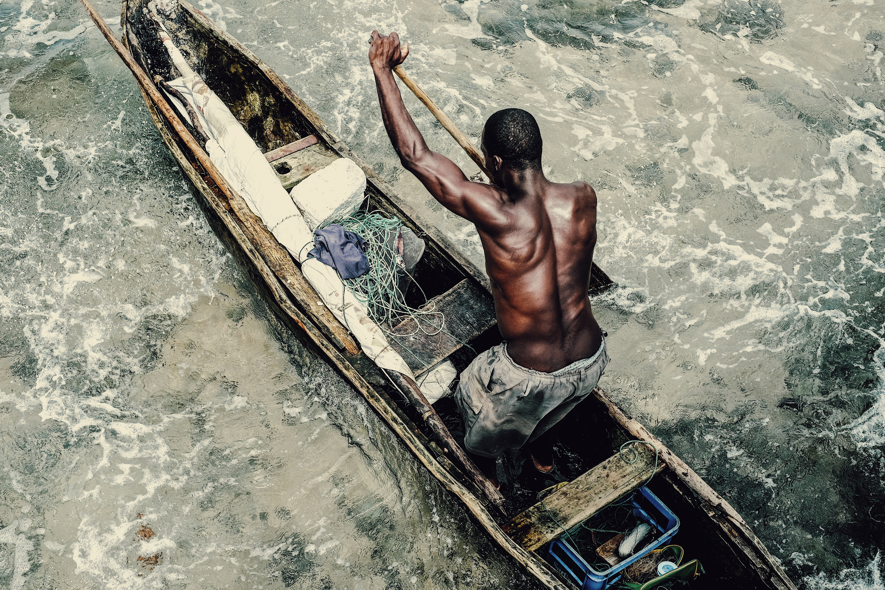 A man paddles his canoe, viewed from above. The muscles on his back are tense. 