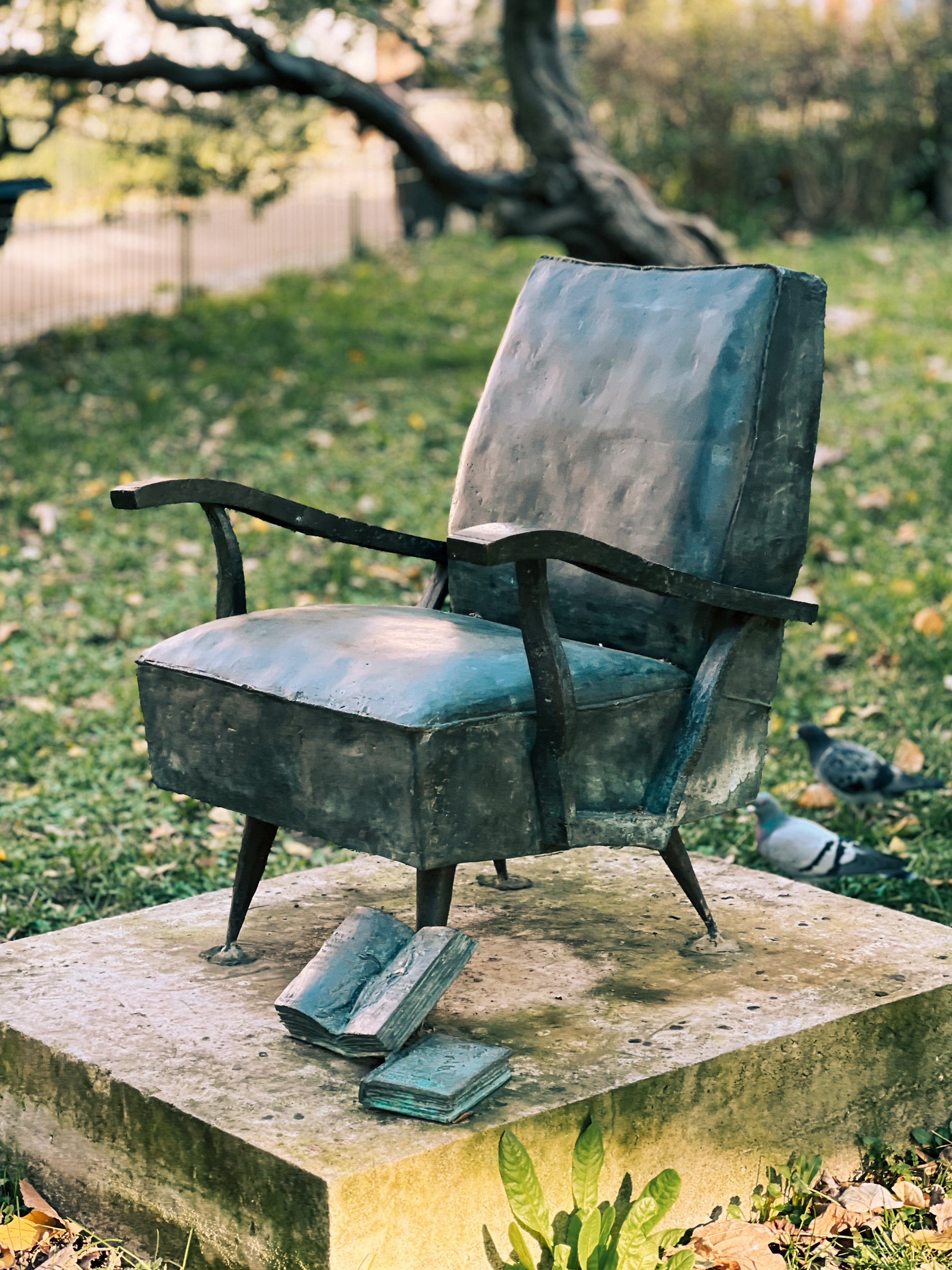 A bronze sculpture of an armchair and books on a stone pedestal, set in a park with green grass and trees in the background. There are pigeons on the ground nearby.