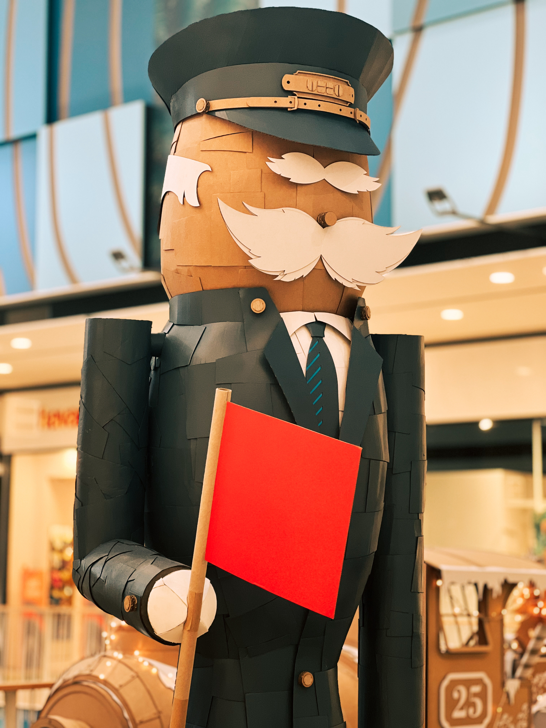 A large, stylized figure made of cardboard resembling a doorman with a mustache, holding a sign, displayed inside a mall setting.
