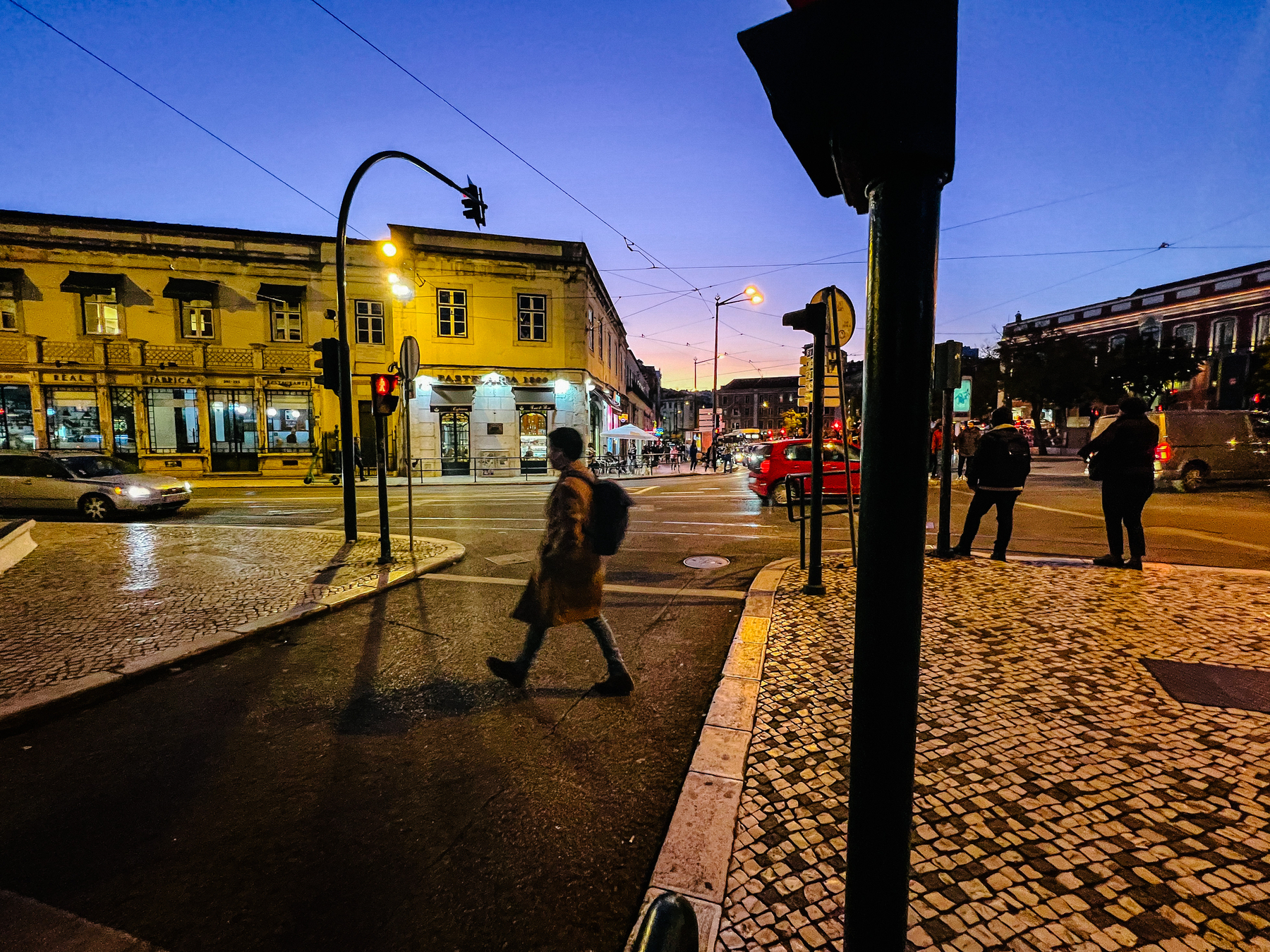 Sunset scene, a person crosses the street while cars drive by, and others wait for the green light. 