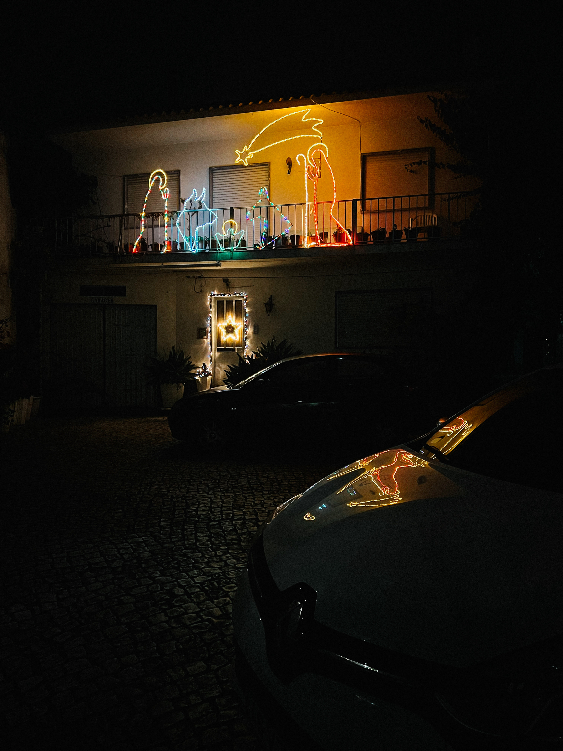 A balcony with Christmas decorations, including illuminated figures of a star and a nativity scene, viewed at night from a street with parked cars.