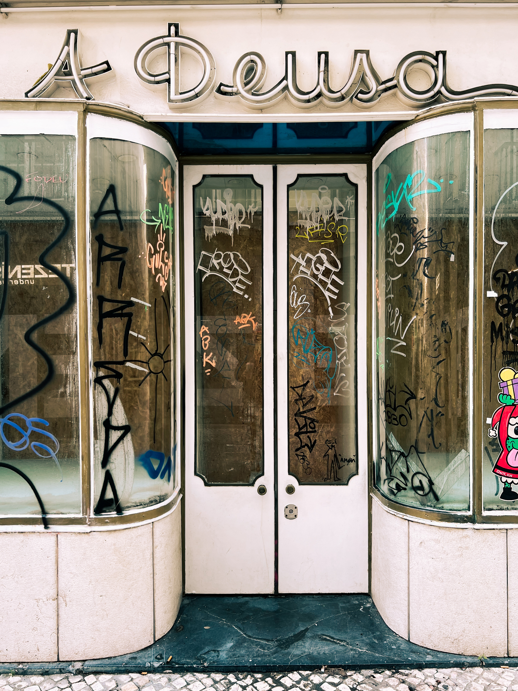 A closed shop, windows graffitied. “A Deusa” (The Godess) still visible above door, in neon. 