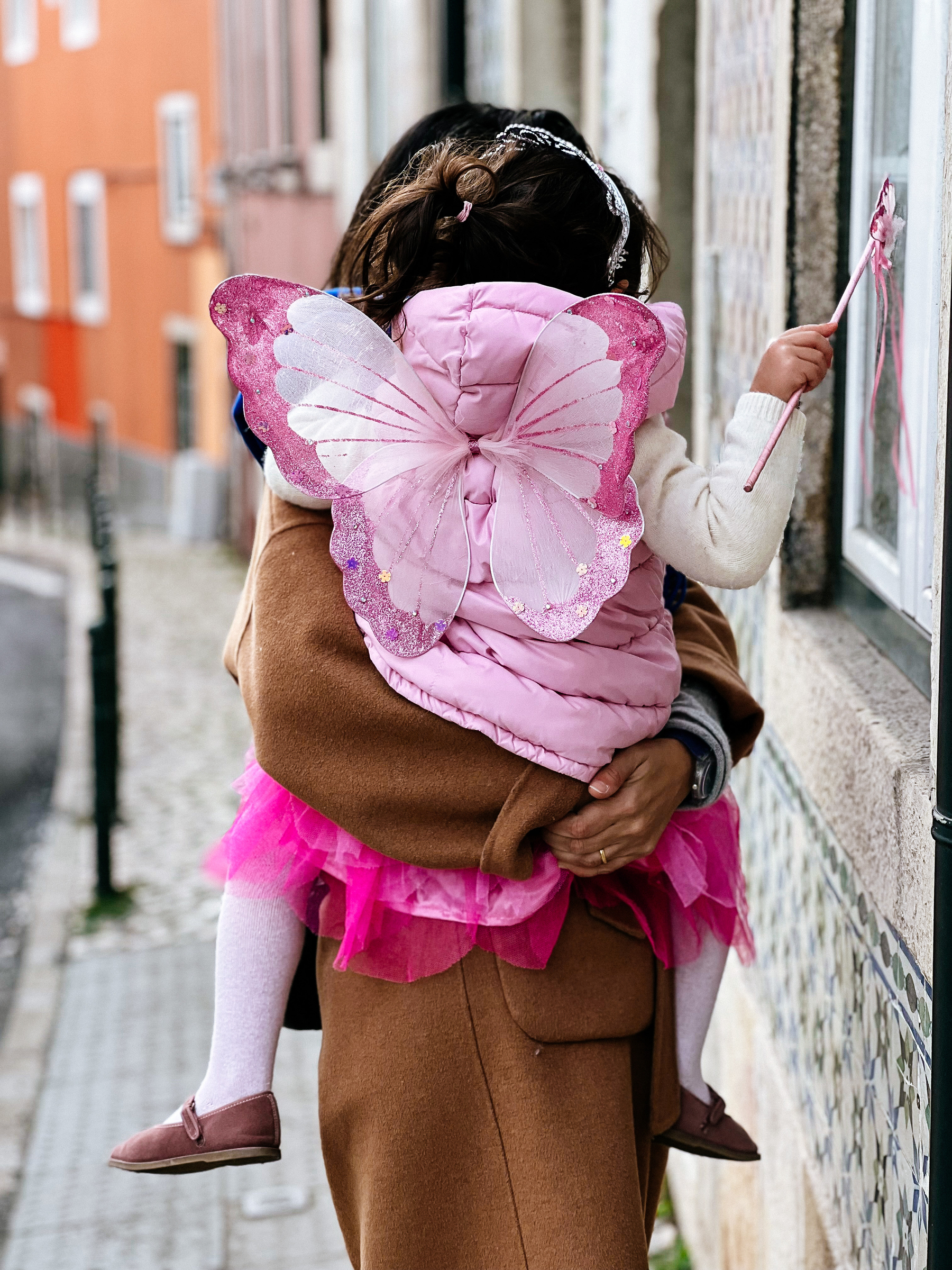 A toddler dressed as a pink fairy taps a window with her magic wand, while her mother holds her. 