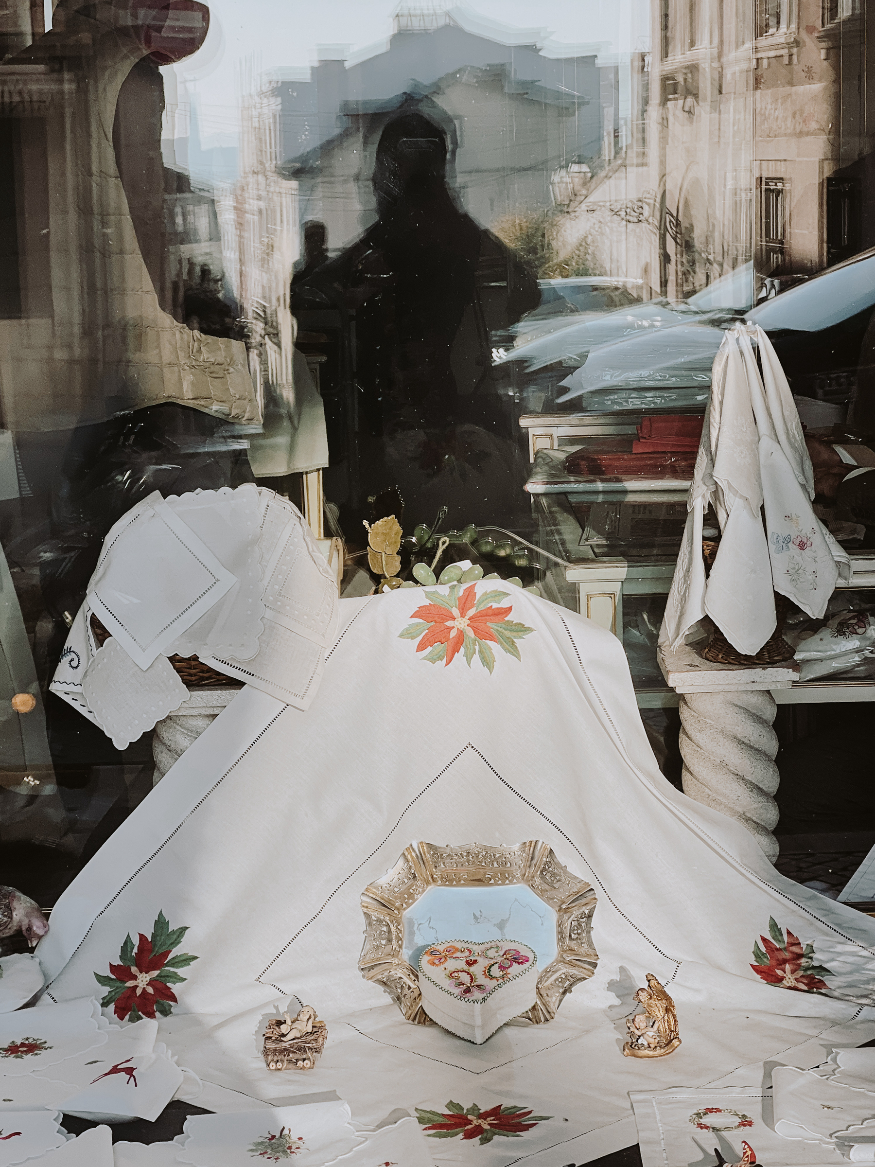A shop window display featuring decorative table linens with embroidered floral designs, alongside a variety of small decorative items. A person&rsquo;s silhouette is reflected in the glass.