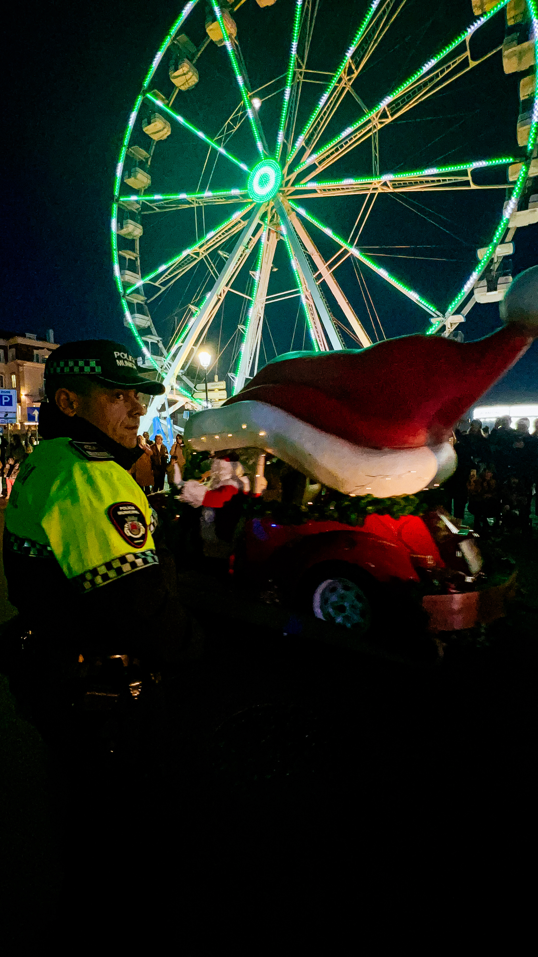 A night scene at a festive event with a ferris wheel illuminated with green lights in the background. In the foreground, a person in a police uniform is visible, and a parade float decorated to look like a whimsical car with a giant Santa Claus